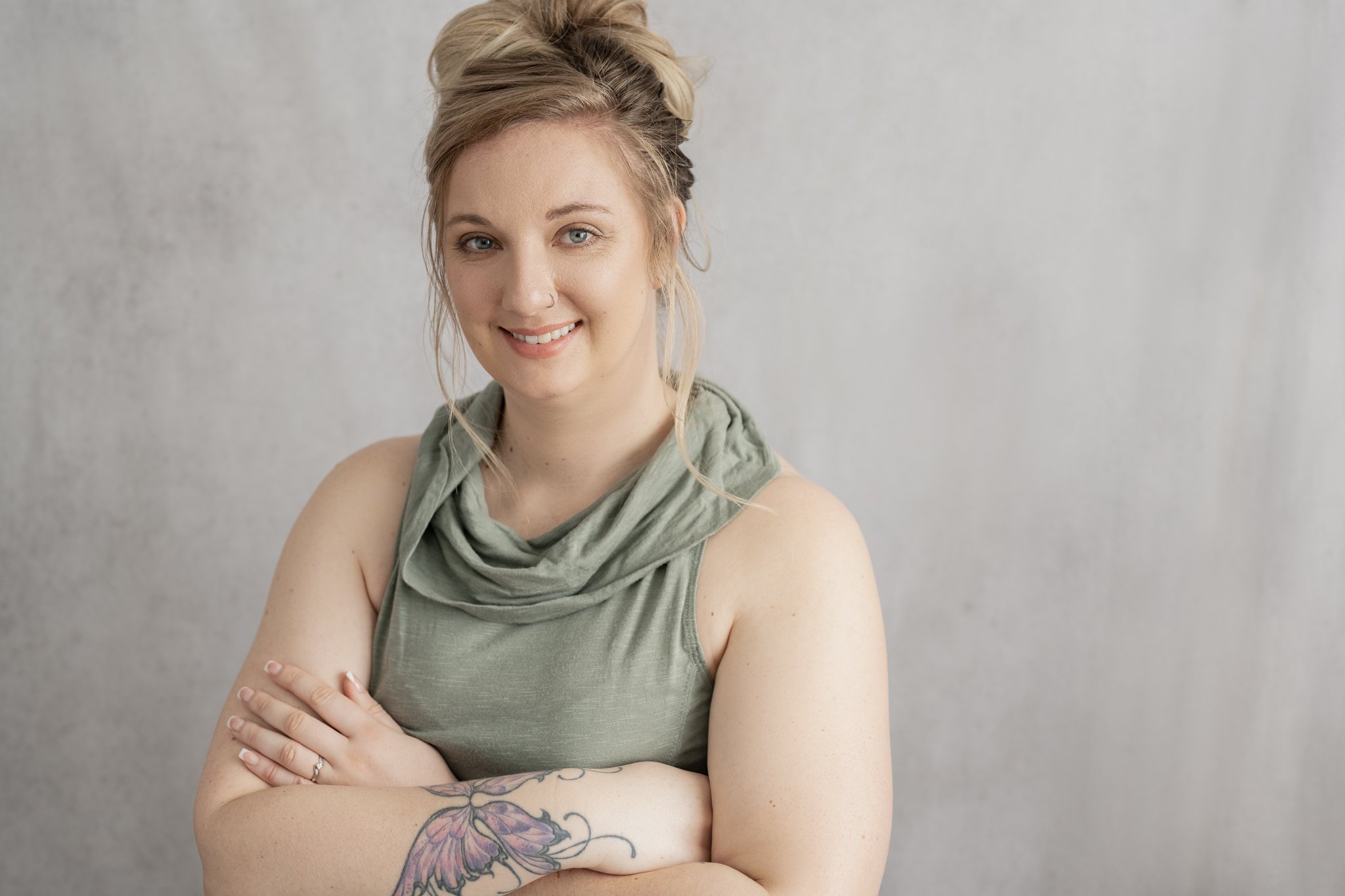 A woman with blonde hair styled in a messy updo, wearing a sleeveless green top with a cowl neckline, standing against a plain light gray background, smiling and crossing her arms, revealing a large purple and black tattoo on her right forearm.