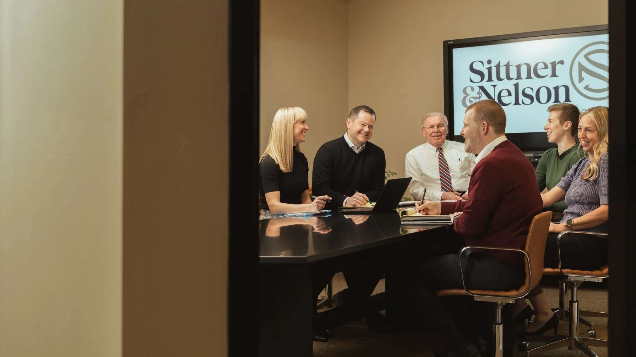 A business meeting with seven people sitting around a conference table, smiling and talking, with a large screen displaying the logo 'Sittner & Nelson' in the background.
