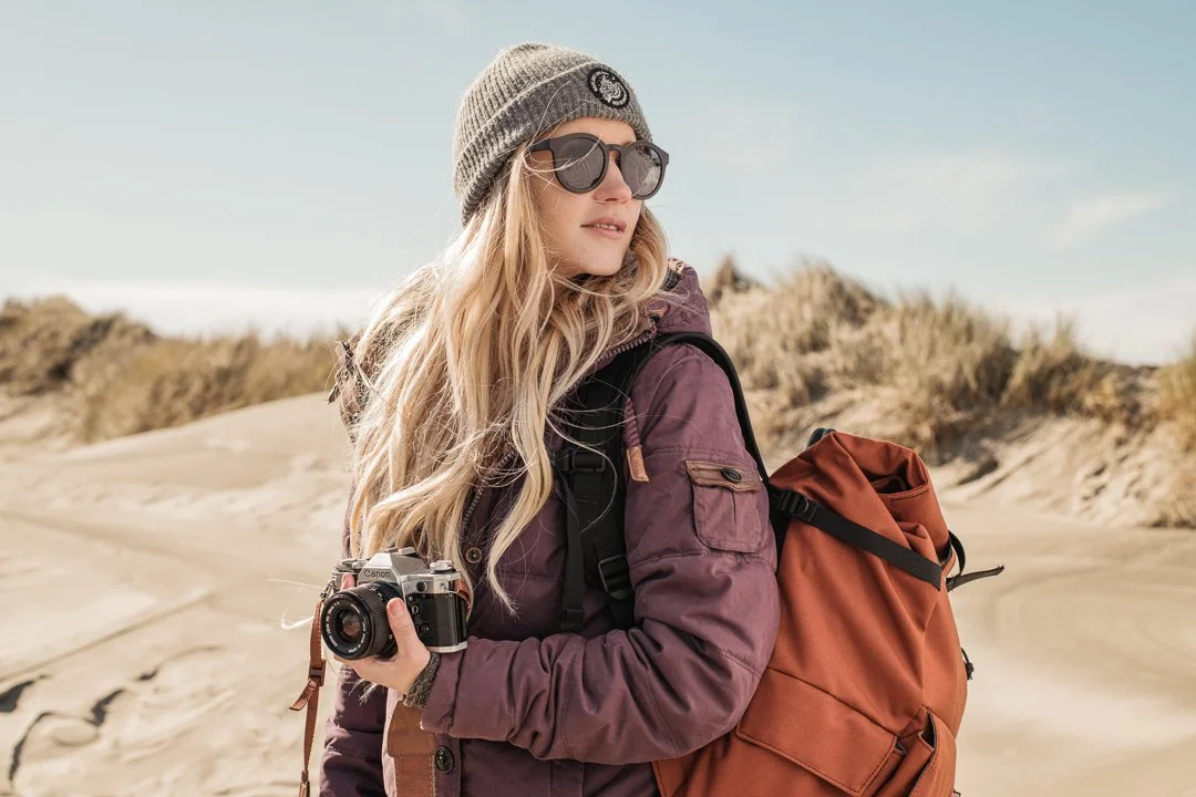 A woman with long blonde hair, wearing sunglasses, a gray beanie, a purple jacket, and a backpack, holding a camera in a sandy outdoor landscape with dunes and sparse vegetation.