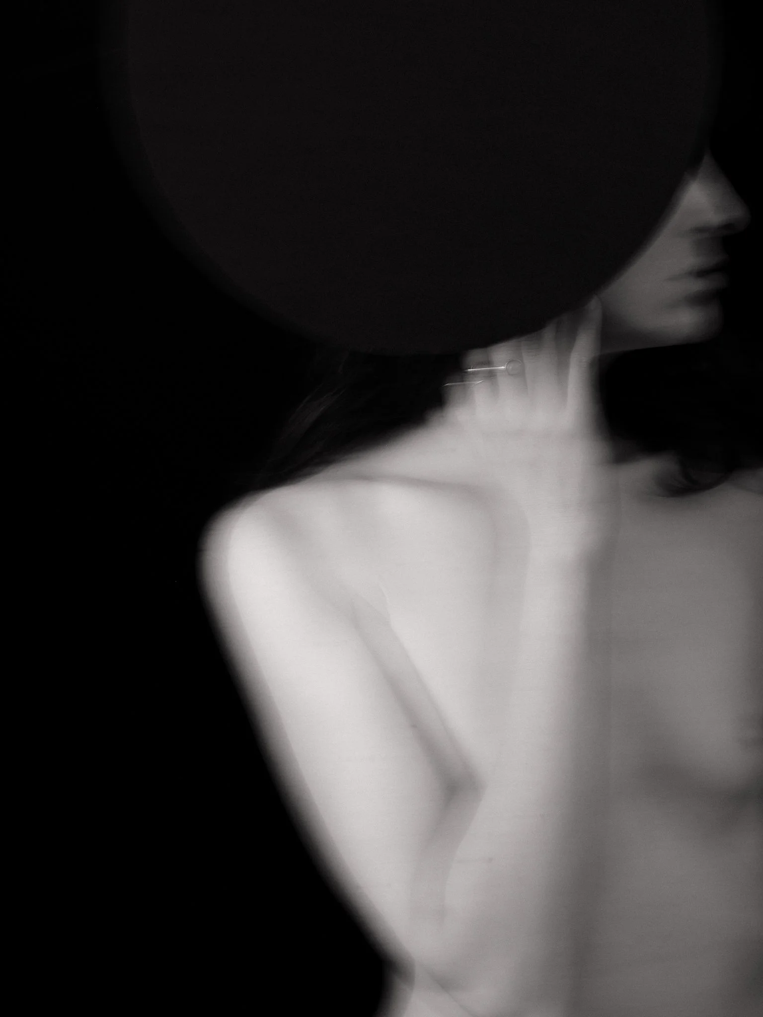 Black and white photo of a woman wearing a black wide-brimmed hat, with her face partially obscured, and her hand resting near her neck.