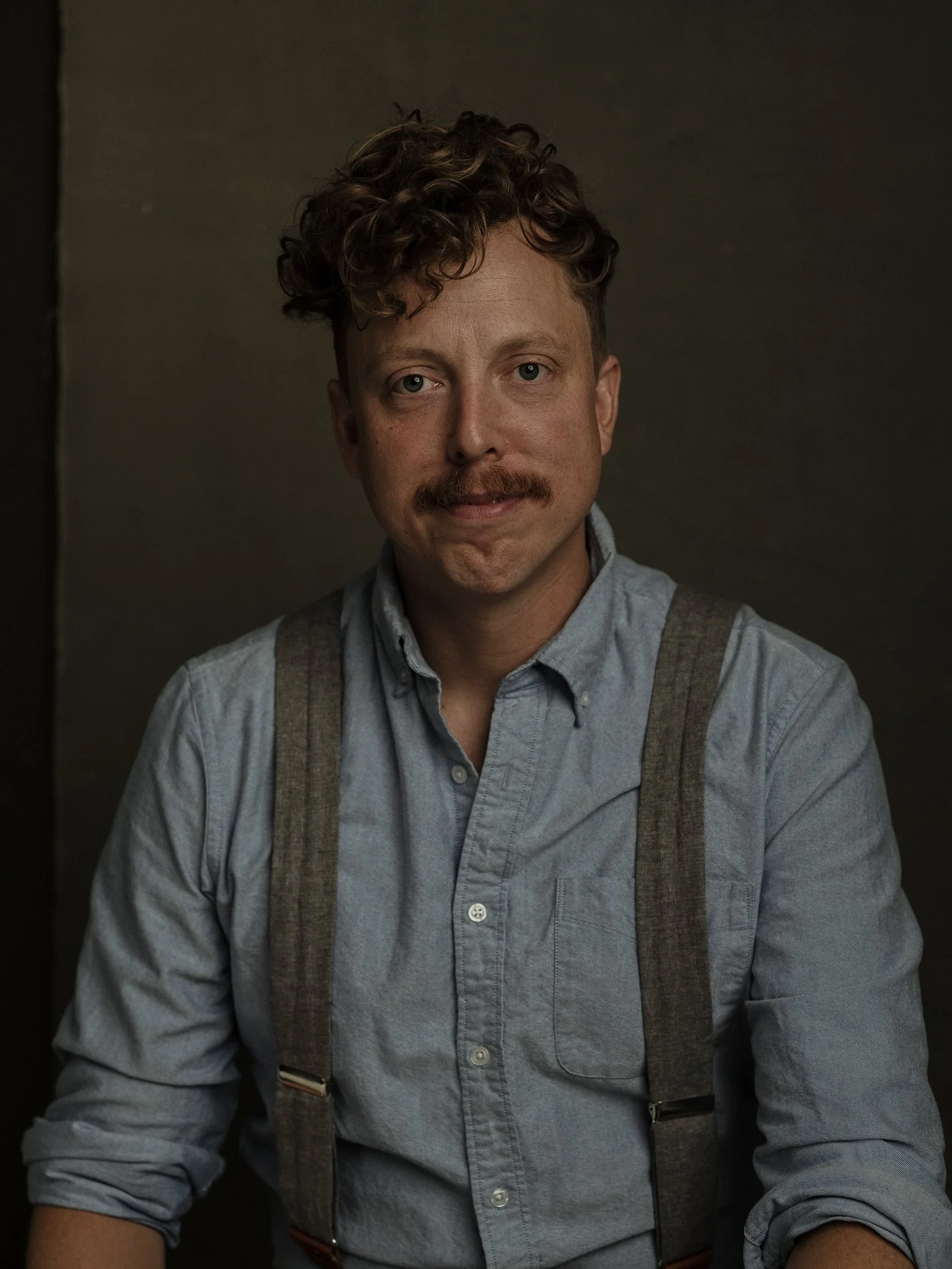 A man with curly brown hair, a mustache, and light skin wearing a light blue shirt with suspenders, looking at the camera against a dark background.