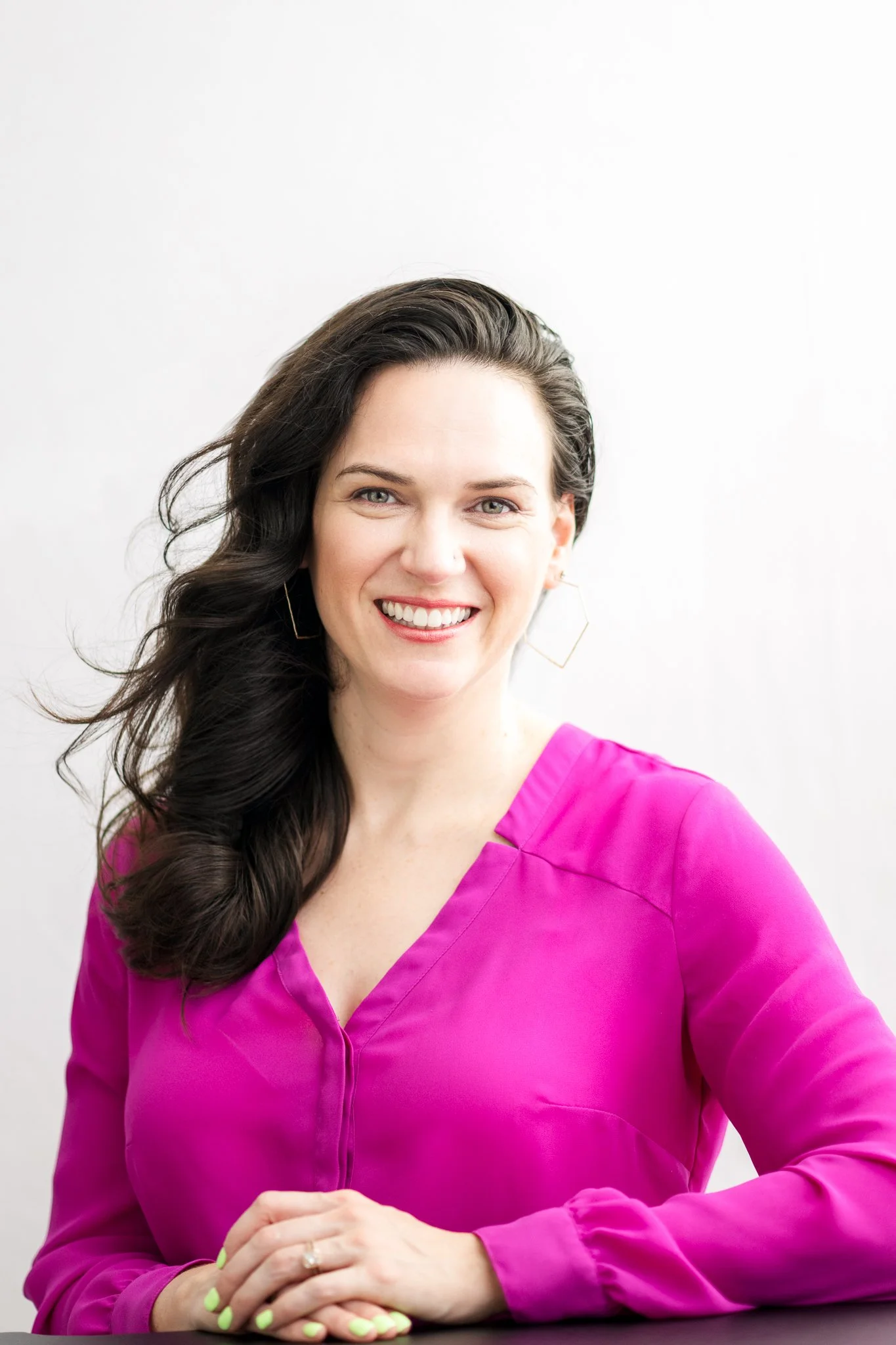 A woman with long, dark wavy hair wearing a bright pink blouse and gold geometric earrings, smiling in front of a white background.
