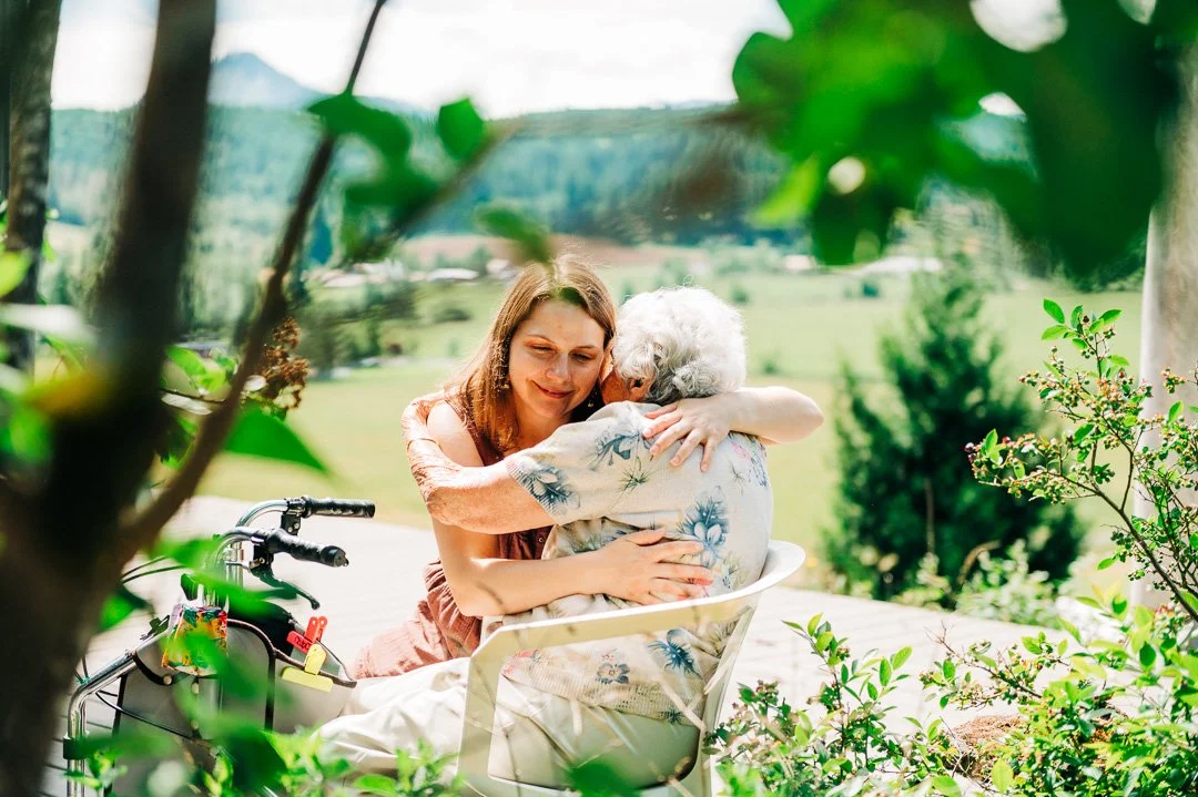 A young woman hugging an elderly woman outdoors with green trees and a field in the background.