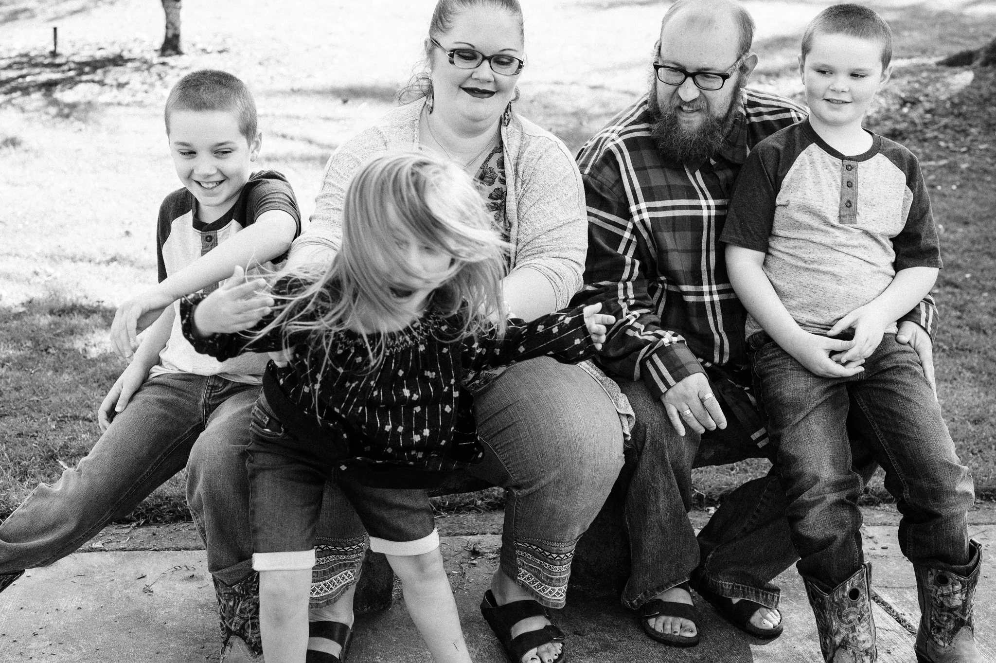 A family of six sitting on a park bench. Two boys, two girls, a woman, and a man, all smiling and playing.