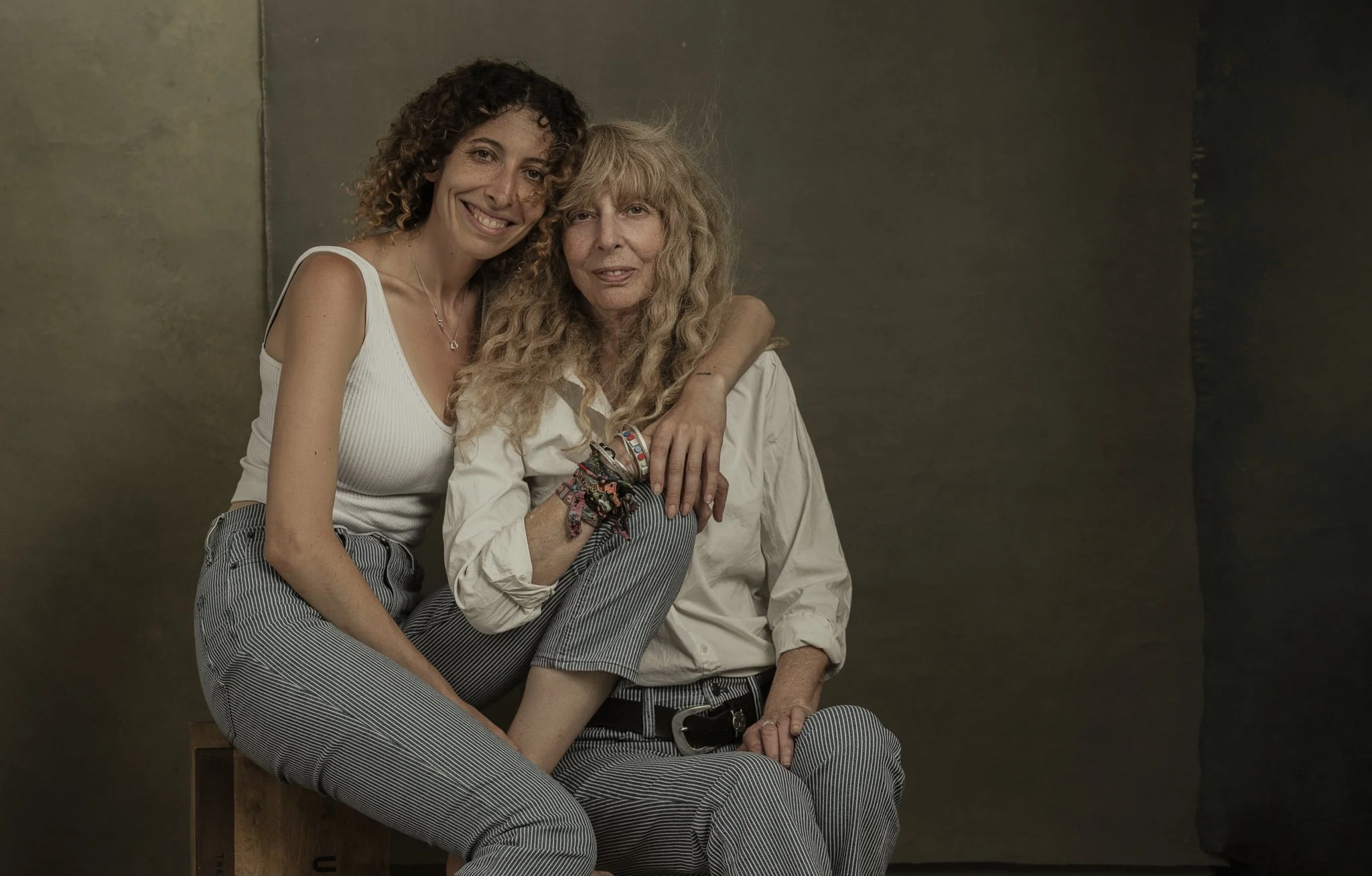Two women sitting together, one younger with curly hair and a white tank top, the other older with wavy blonde hair and a white shirt, with her arm around the younger woman's shoulder.