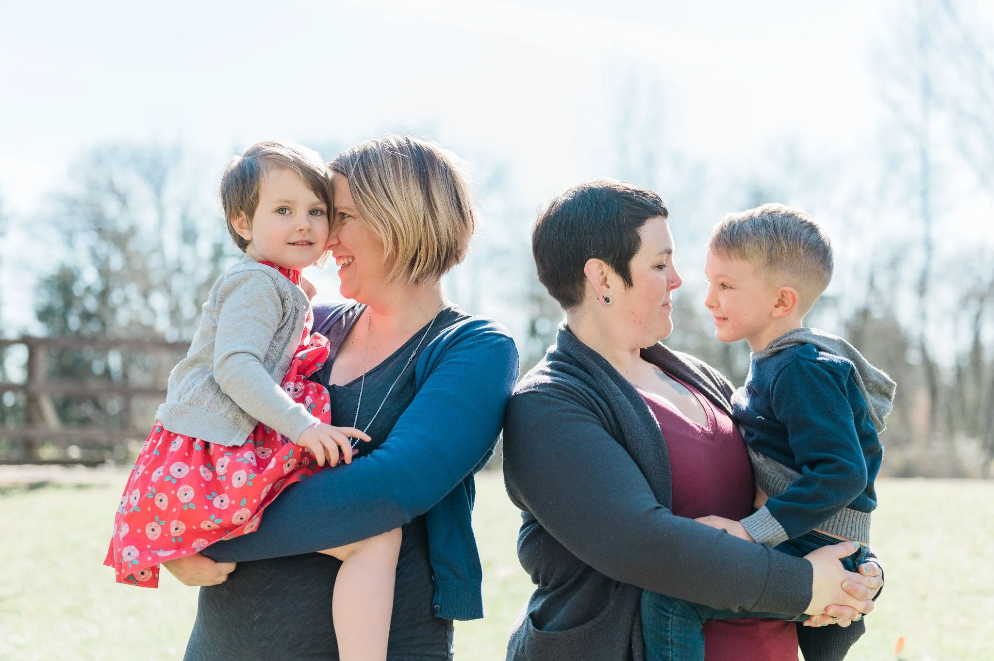Two women, each holding a child, standing outside on a sunny day with trees in the background.