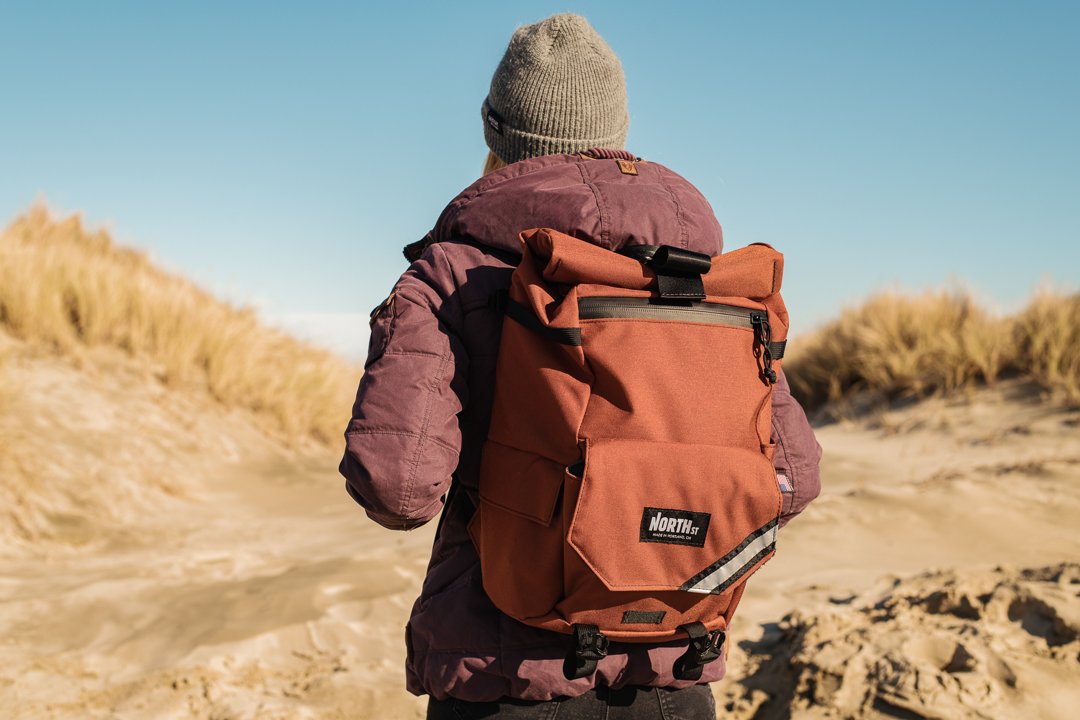 Person with a large orange backpack walking through sandy dunes with grass, wearing a gray beanie and maroon jacket.