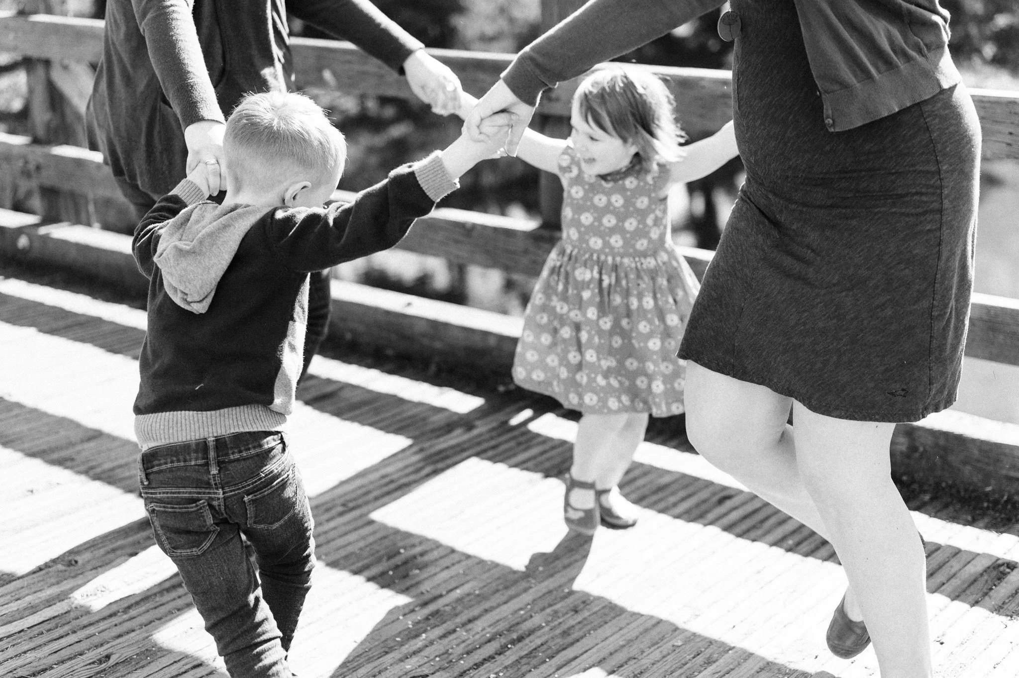 Two children, a boy and a girl, holding hands and dancing on a wooden bridge, with two adults also holding their hands and guiding them.