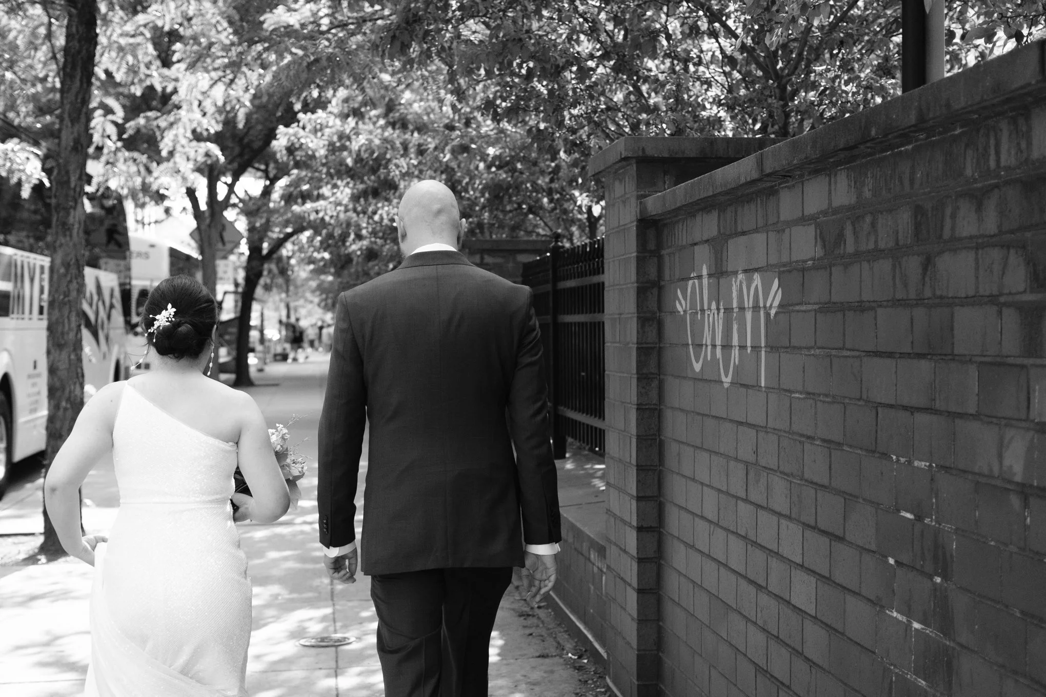 A black and white photograph of a bride and groom walking together down a city sidewalk seen from behind. The bride wears a one-shoulder wedding gown with her hair styled in an updo decorated with flowers. The groom is in a dark suit, and they are wa