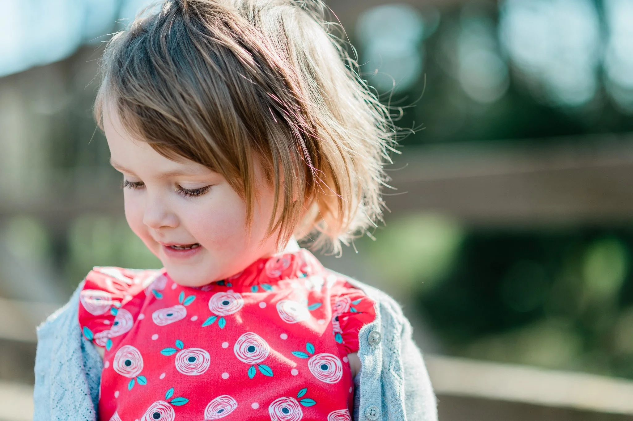 A young girl with short, wavy brown hair smiles while looking down. She is outdoors, wearing a red floral-patterned bib over a light gray jacket.