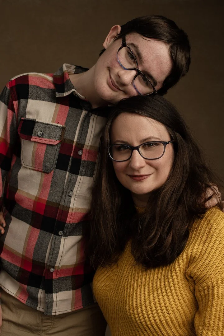 A young man and woman wearing glasses, smiling and posing closely together in a studio portrait.
