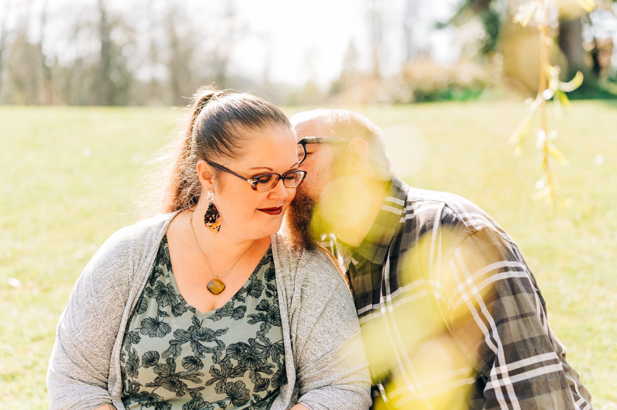 A woman with glasses and earrings, dressed in a floral top and cardigan, leaning her head towards a bearded man with glasses in plaid shirt, in an outdoor park setting, with sunlight and blurred yellow leaves in the foreground.