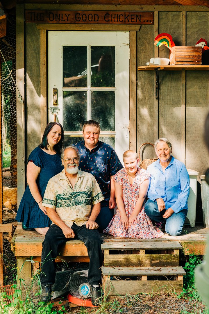 A group of five people, including three women, one man, and one girl, poses together on a wooden porch outside a rustic building. They are smiling and appear to be enjoying themselves. The sign above the door reads 'The Only Good Chicken...' and ther