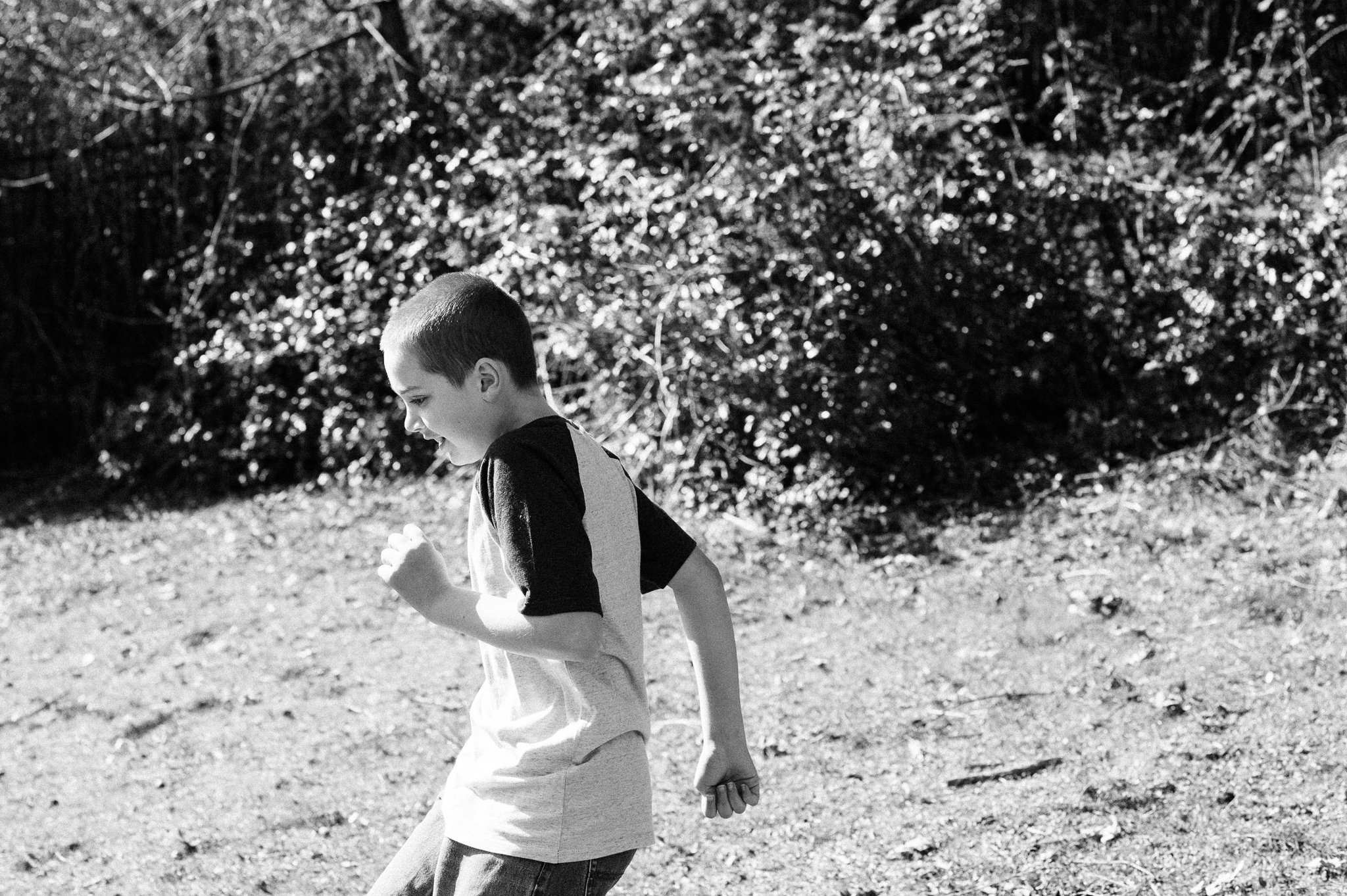 A young boy plays outdoors, running on a dirt path with a big smile, surrounded by trees and bushes.