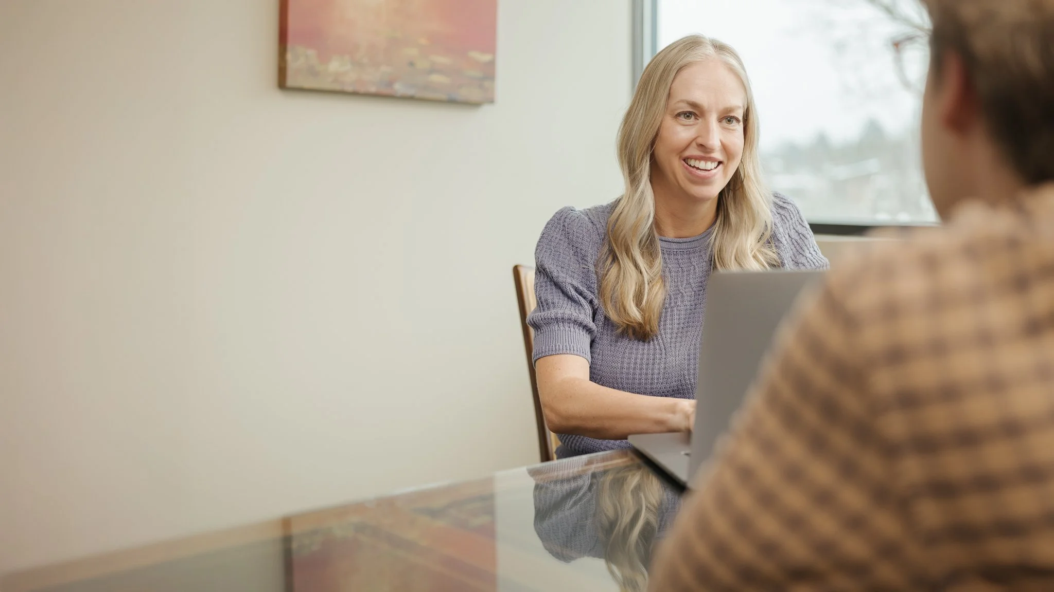A woman with long blonde hair smiling and talking at a table with another person, and a laptop open in front of her. The background features a window and a piece of abstract art on the wall.