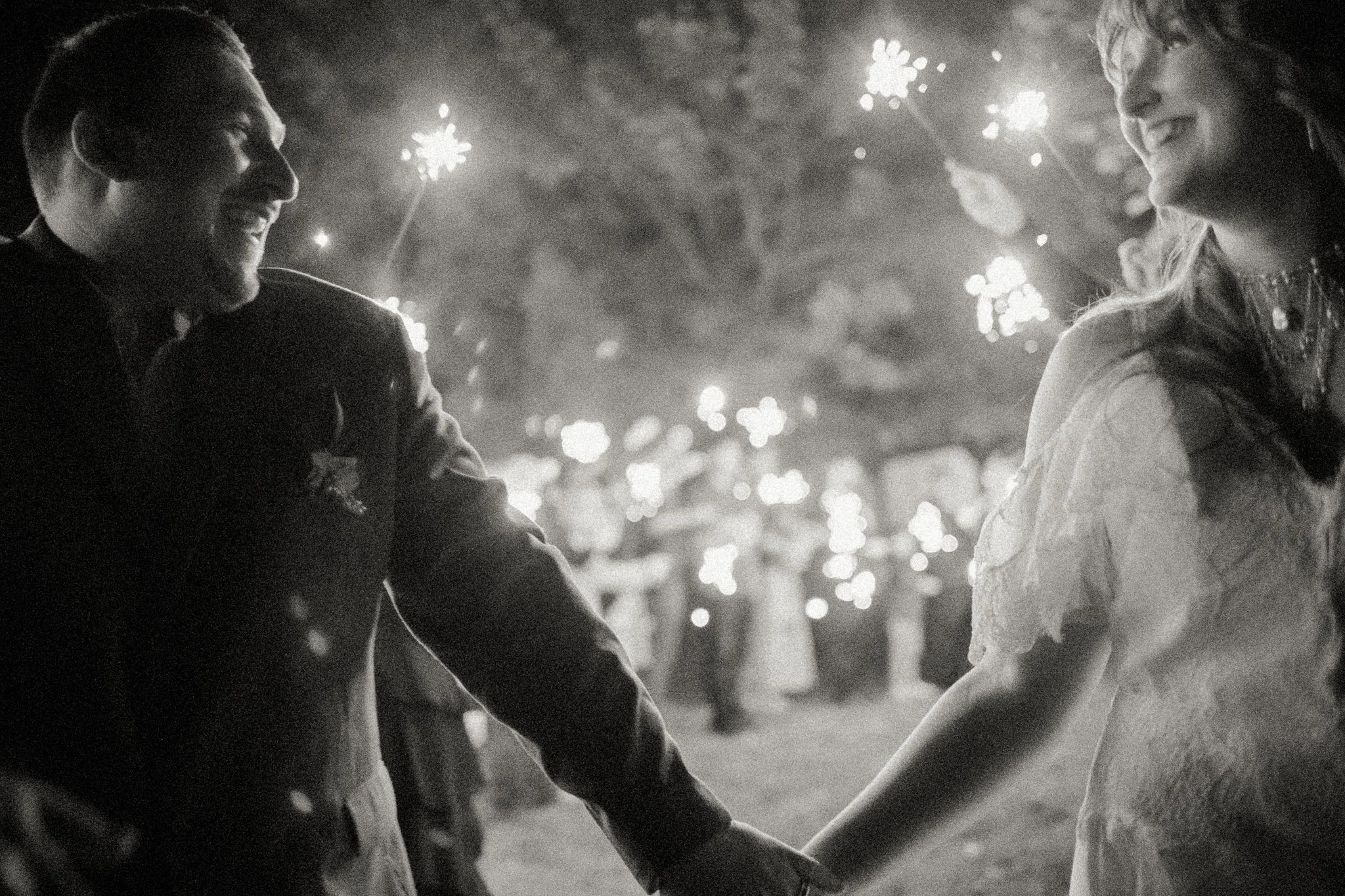 Two couples holding hands and smiling at each other while fireworks light up the night sky.