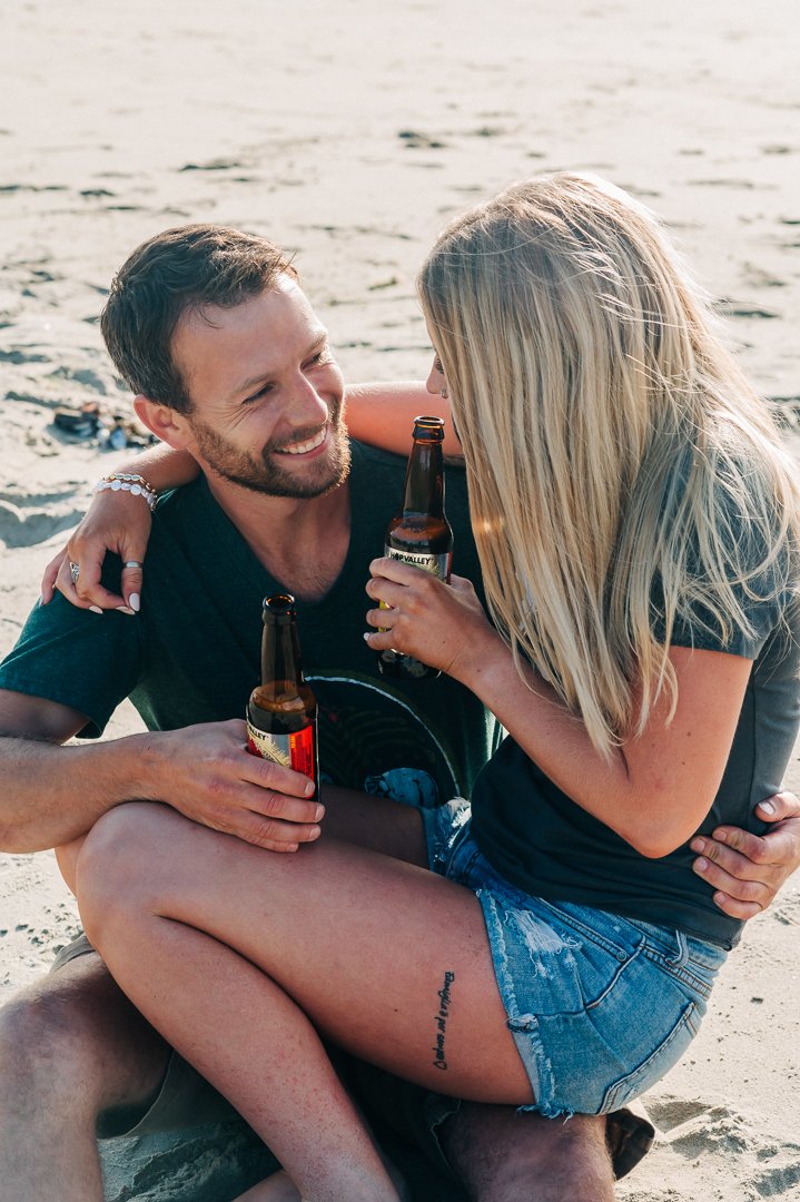 A couple sitting on the sand at the beach, smiling and talking while holding bottles of beer.
