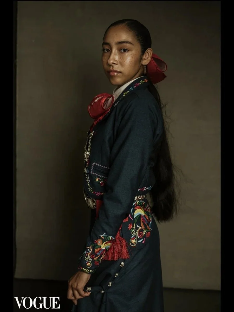 A young woman with long dark hair, wearing traditional embroidered clothing with red accents, stands against a plain background, looking ahead with a neutral expression.