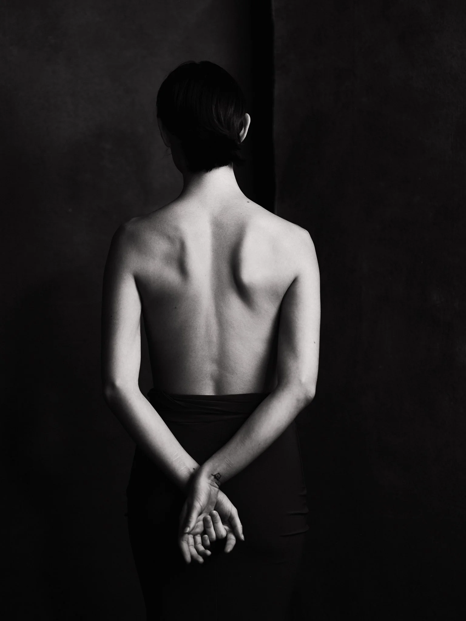 Back view of a person with short dark hair, topless, standing with hands clasped behind back in a dimly lit, black-and-white setting.