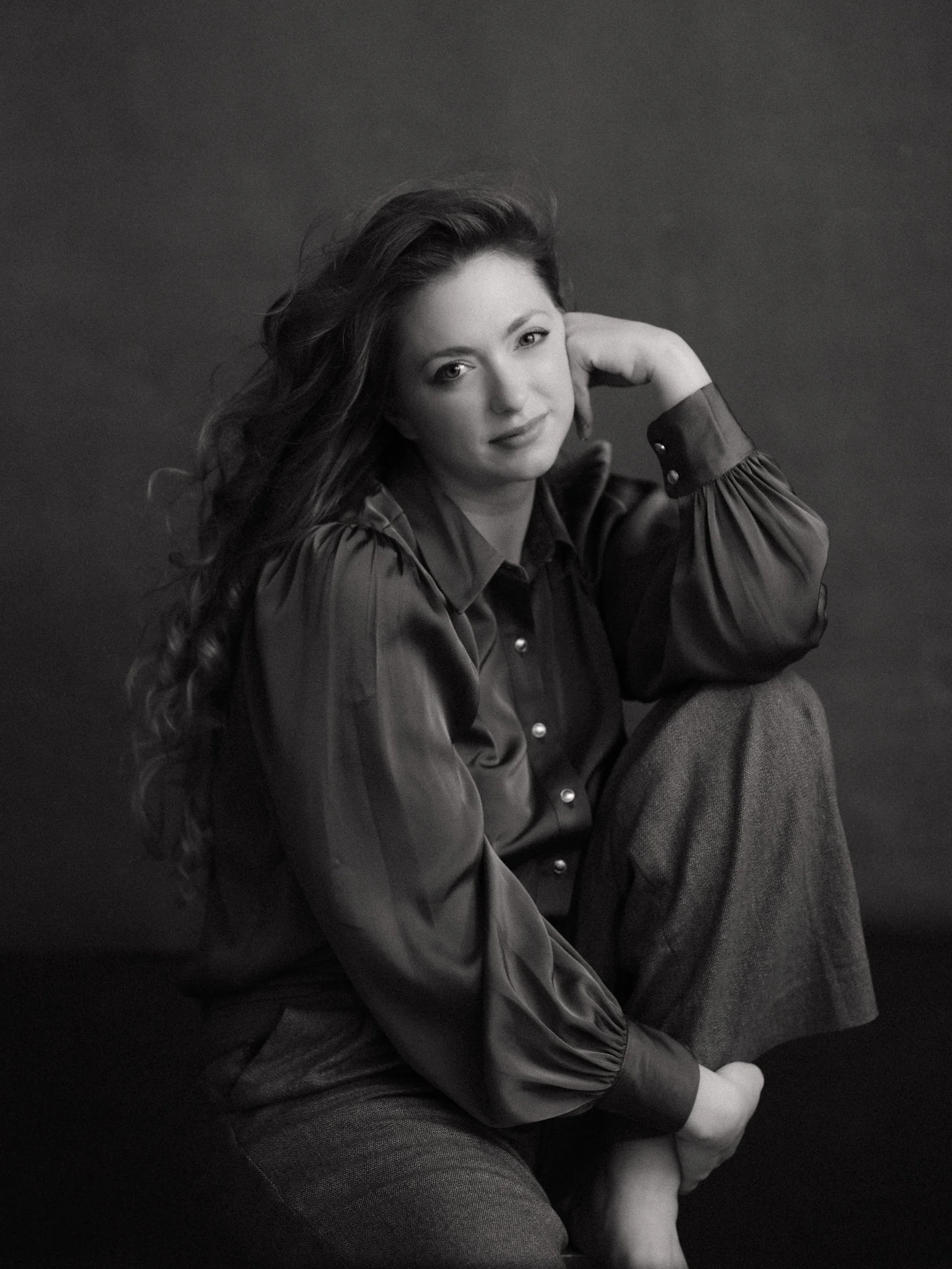 Black and white portrait of a woman with long wavy hair, sitting with one hand resting on her head. She is wearing a satin shirt and loose trousers, looking at the camera with a slight smile.