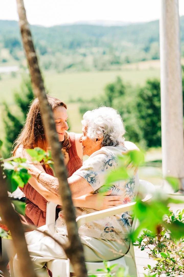 A young woman and an elderly woman sharing an embrace outdoors with a forested landscape in the background.
