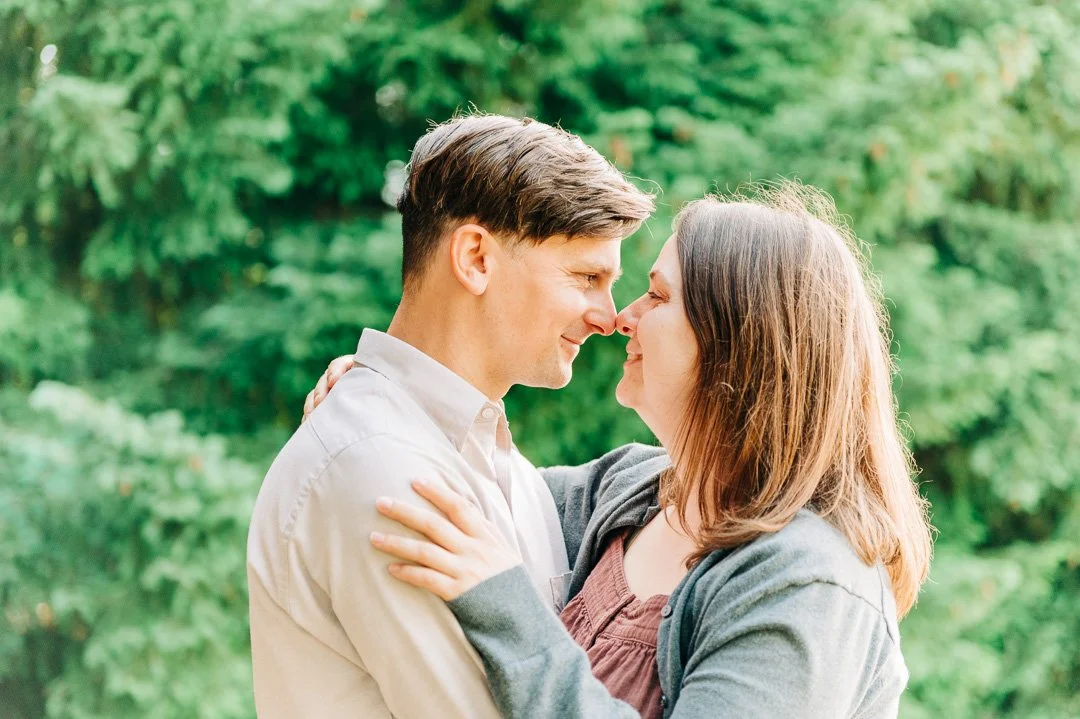 A couple in love touching noses and smiling outdoors in front of green trees.