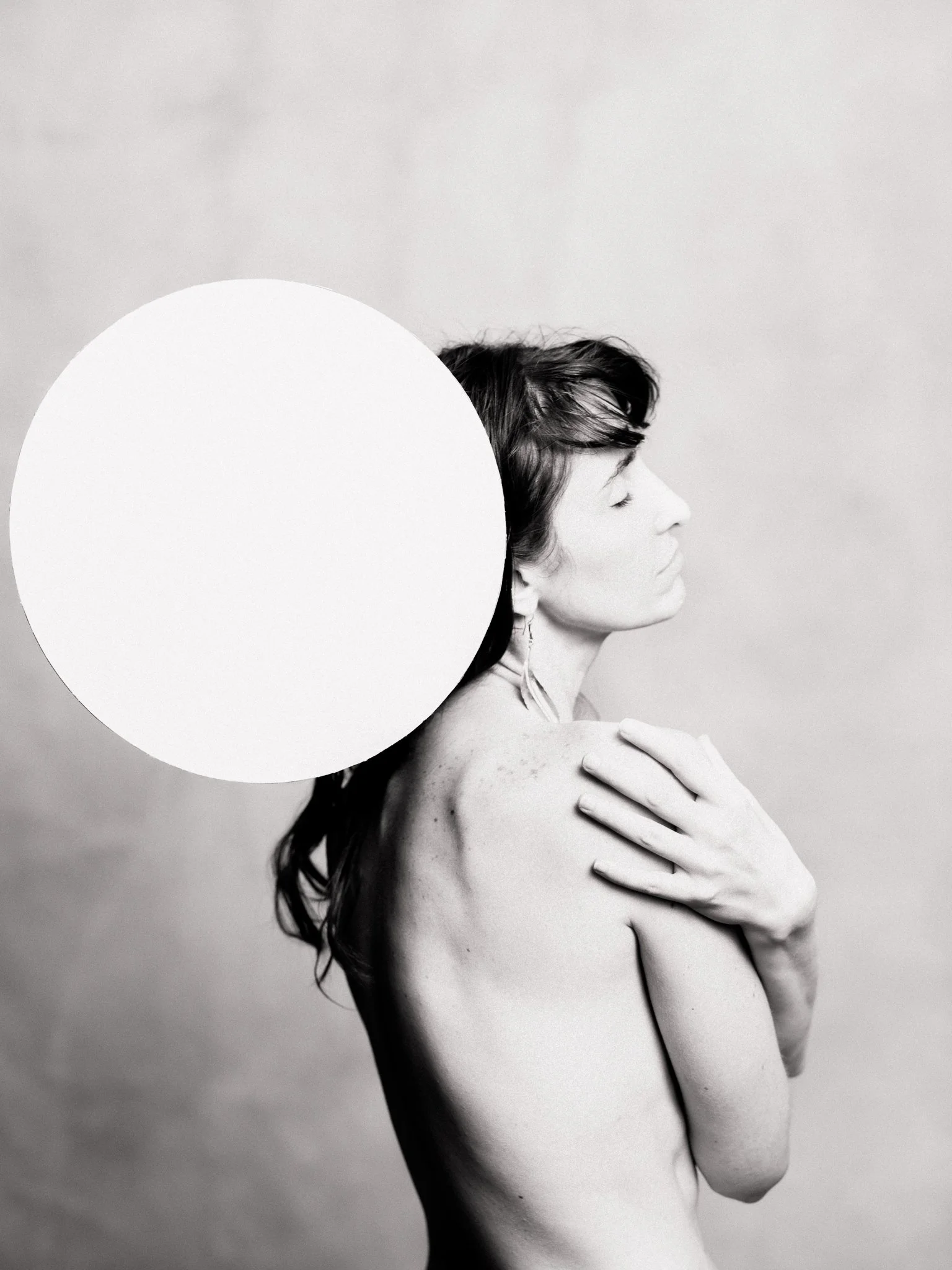 Black and white photograph of a woman with short dark hair, eyes closed, standing with her right hand on her shoulder. A large white circle partially covers her head and upper body against a plain background.