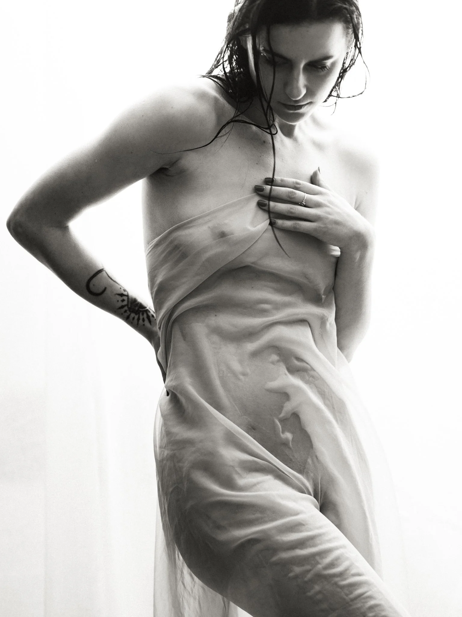 A black-and-white photograph of a woman with wet hair, wearing a translucent cloth that drapes over her body, with one hand covering her chest and looking downward.