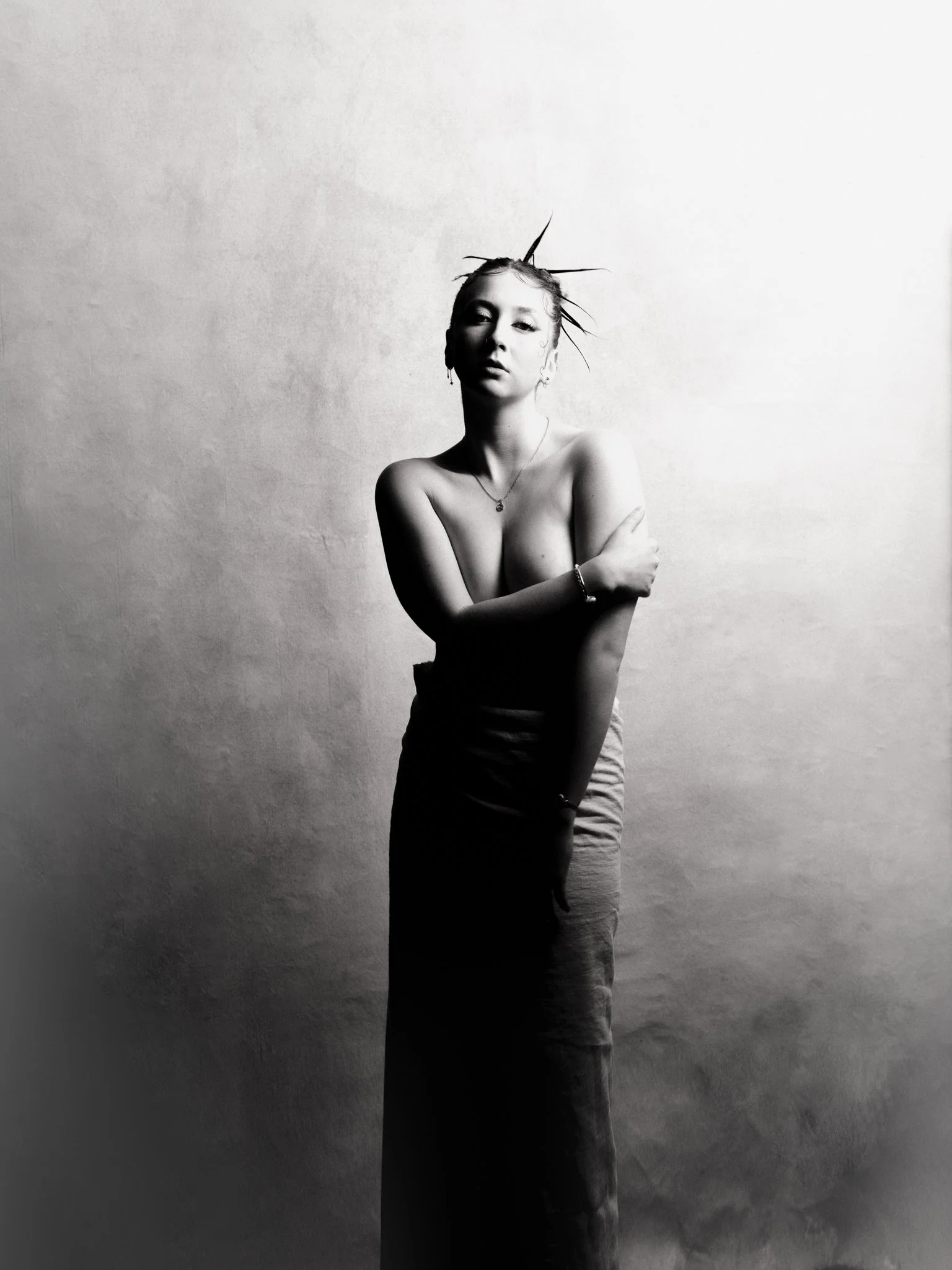 Black and white photo of a topless woman with crossed arms, wearing a long skirt, earrings, a necklace, and a bracelet, standing against a plain wall with styled hair and a serious expression.