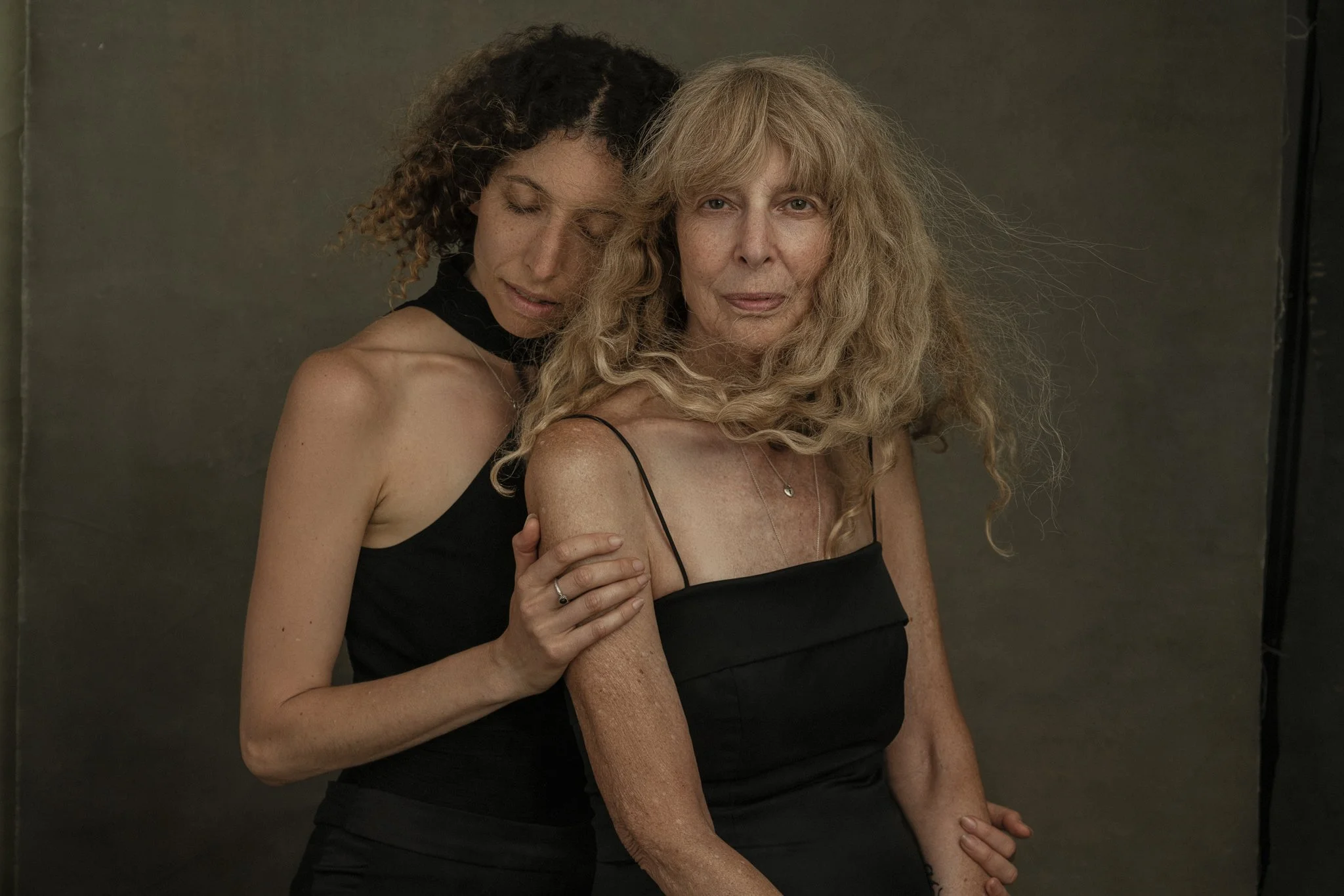 Two women, one with curly dark hair and the other with long blonde hair, are posing together, both wearing black clothing. The woman with dark hair has her eyes closed and is resting her head on the shoulder of the blonde woman, who is looking at the camera with a neutral expression. They are standing against a neutral backdrop.