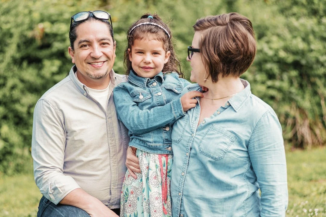 Family of three outdoors: a man, woman, and young girl smiling and enjoying each other's company on a grassy area with trees in the background.