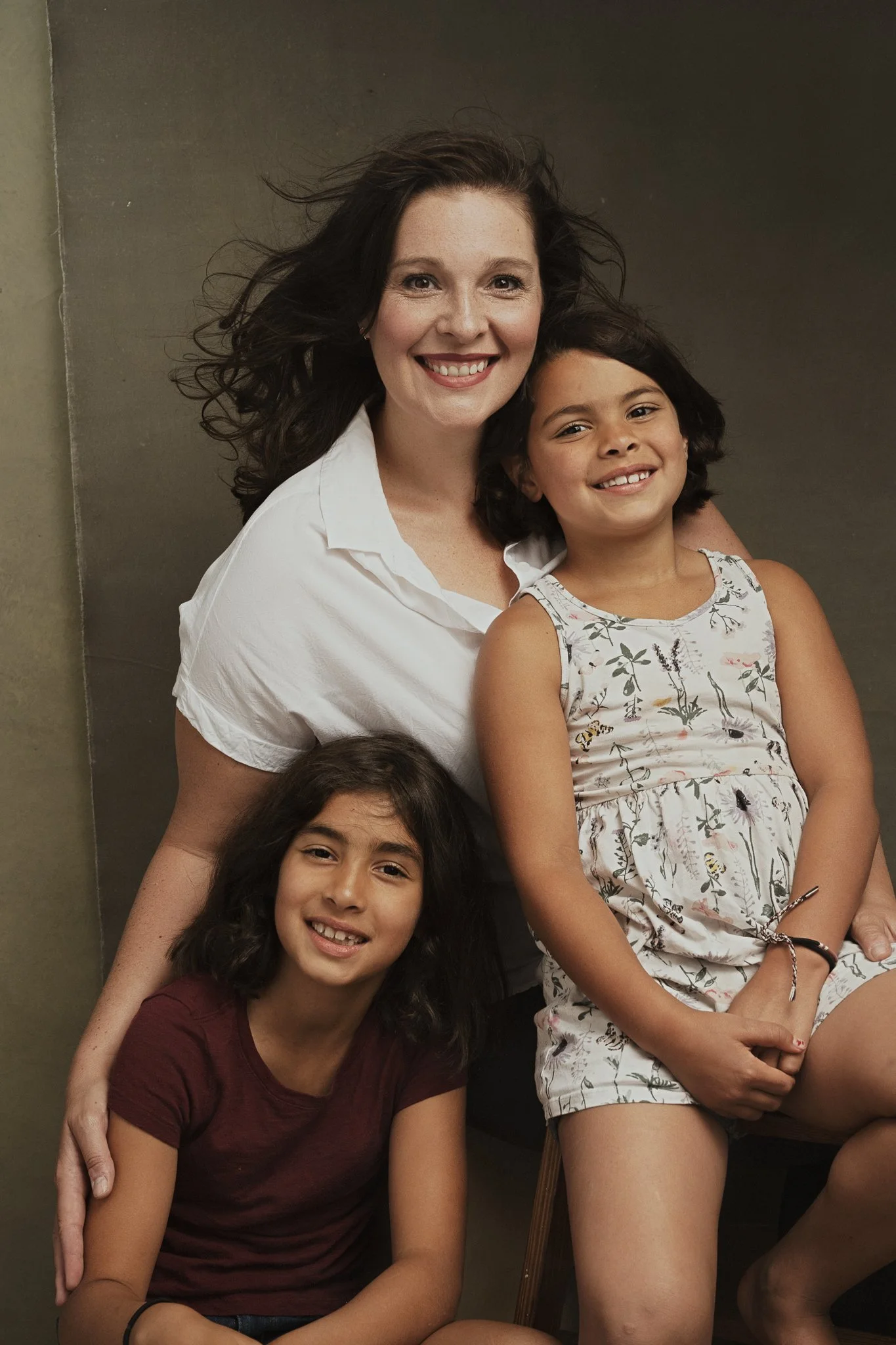 A woman with dark curly hair smiling, seated with two young girls, one with dark curly hair and a floral dress, the other with straight dark hair and a maroon shirt, all smiling.
