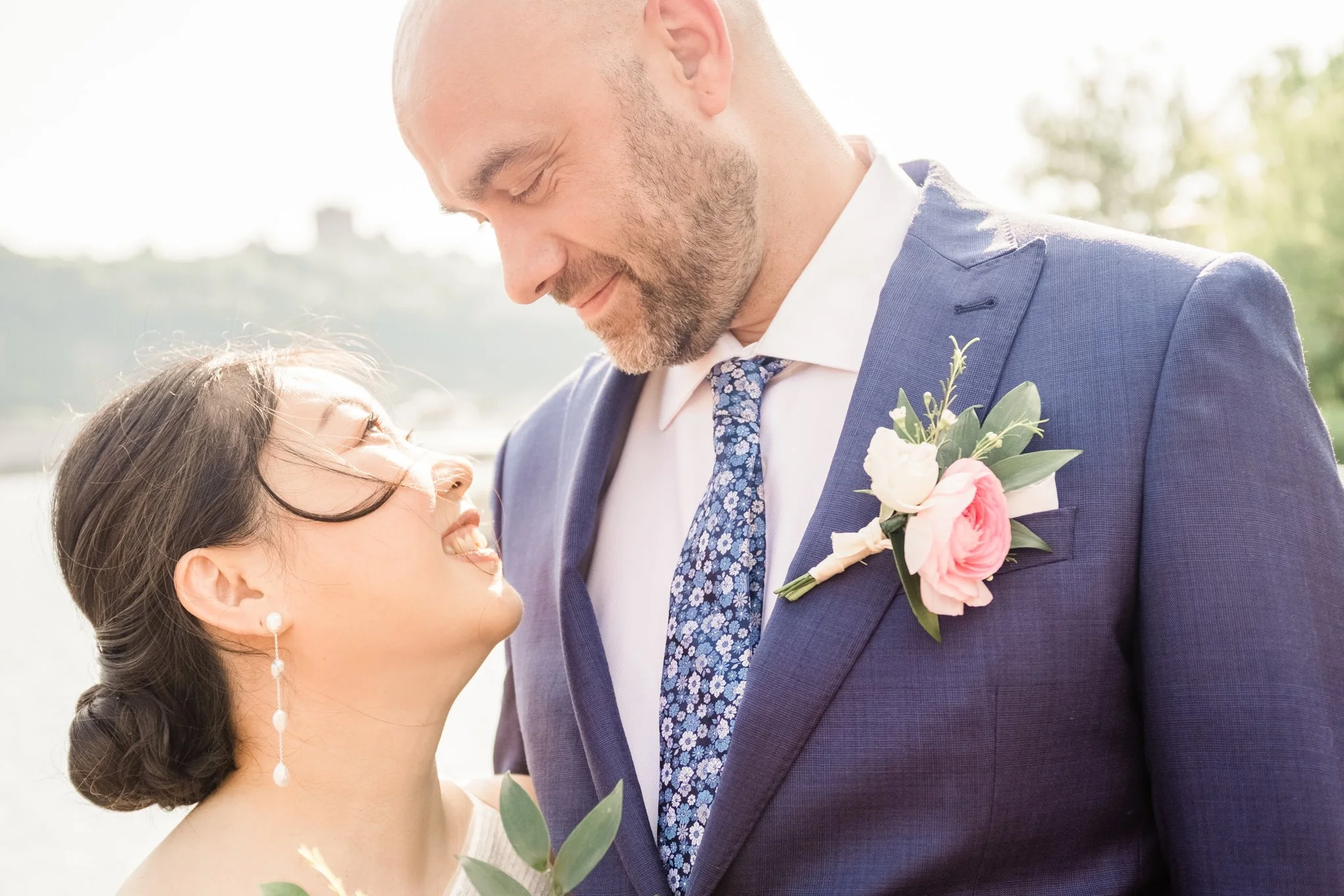 A close-up of a smiling couple dressed in formal attire, standing outdoors with a blurred natural background, sharing an intimate moment.