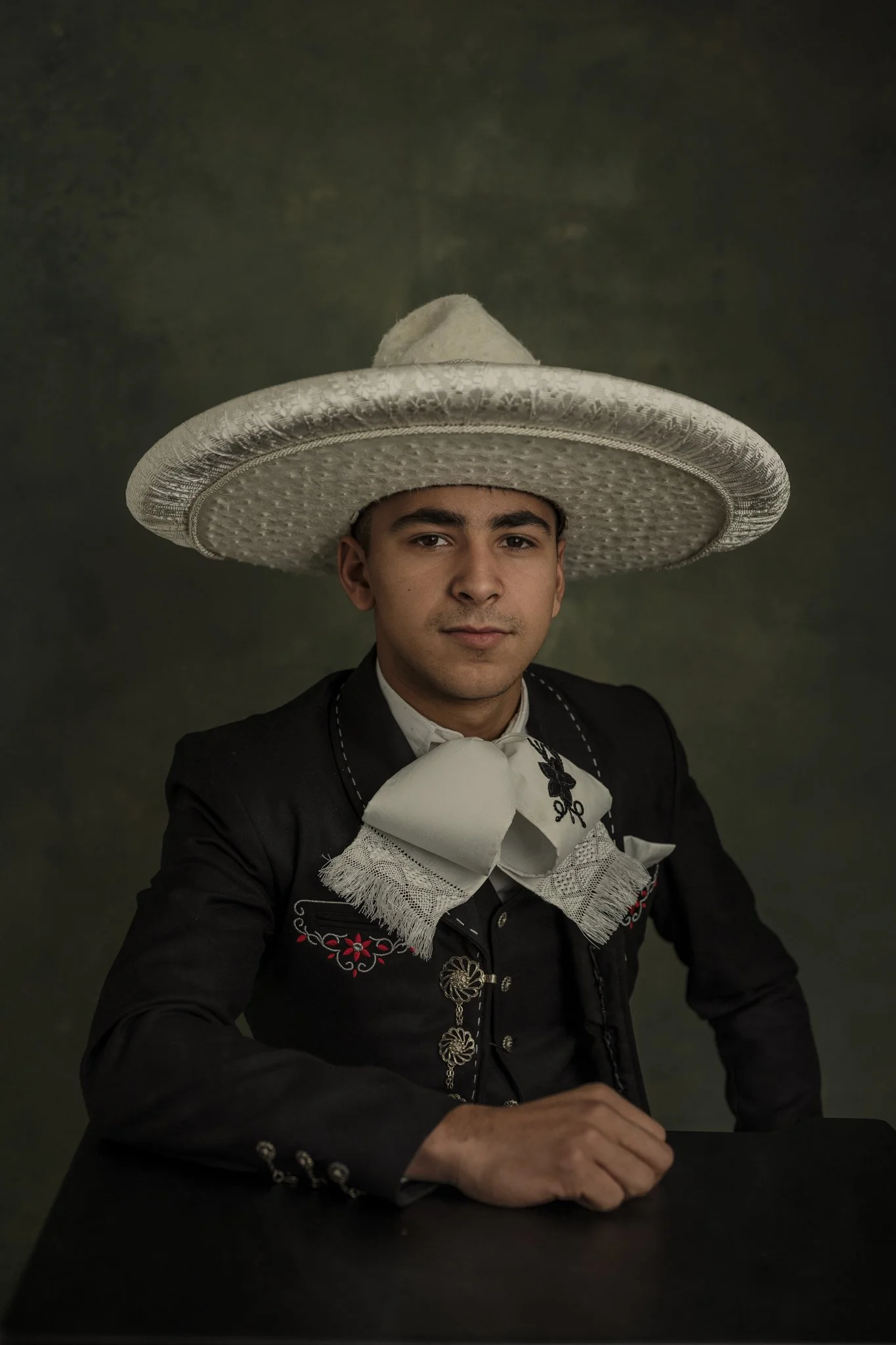 Young man dressed as a traditional Mexican mariachi musician, wearing a large sombrero, black charro suit with embroidered details, and a white bow tie, posing against a dark background.