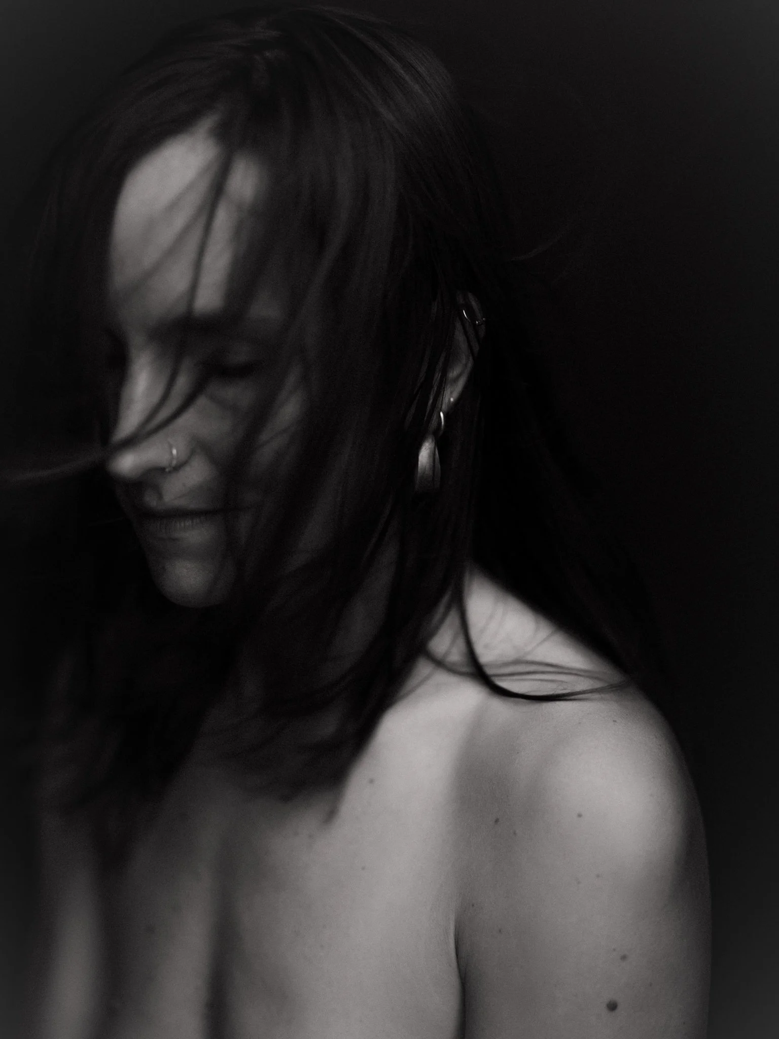 A black and white close-up photo of a woman with wet hair partially covering her face, smiling softly, with her shoulder visible against a dark background.