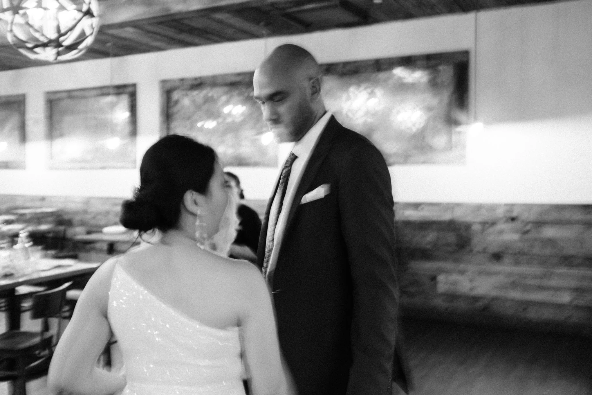 A black and white photo of a man and woman standing close together in a restaurant, with the man looking down at the woman. The woman has dark hair styled in a bun and is wearing an off-the-shoulder dress. The man is dressed in a suit and tie. The ba