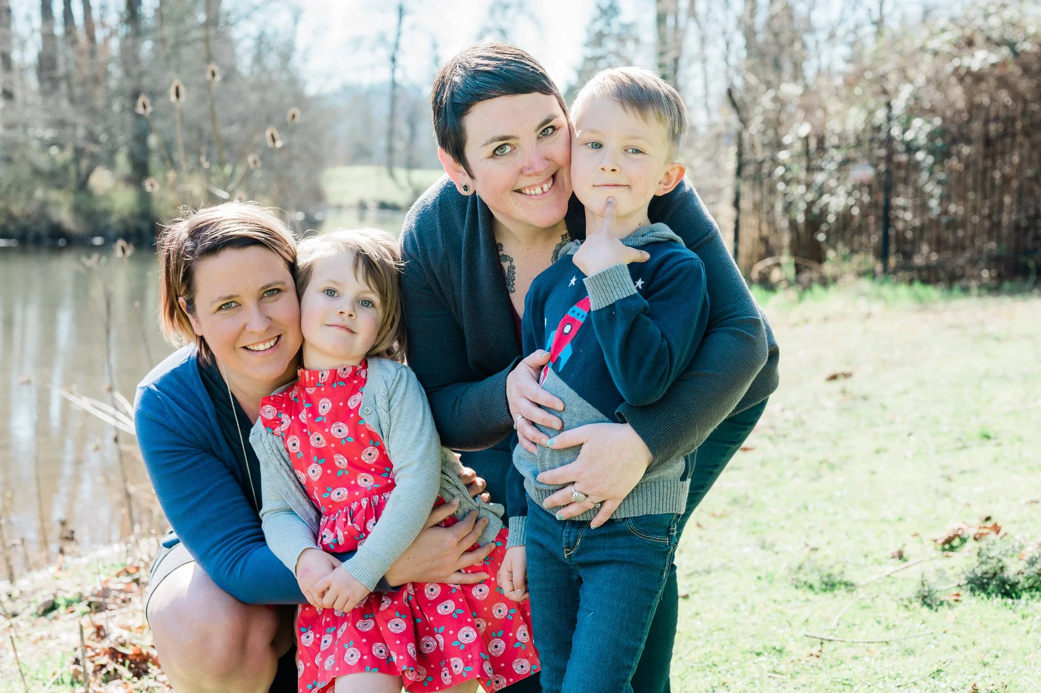 Two women and two children outside near a body of water, smiling and embracing during daytime.