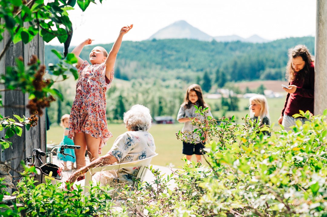 Children playing outdoors with an elderly woman sitting on a chair, surrounded by green plants and a scenic mountain background.