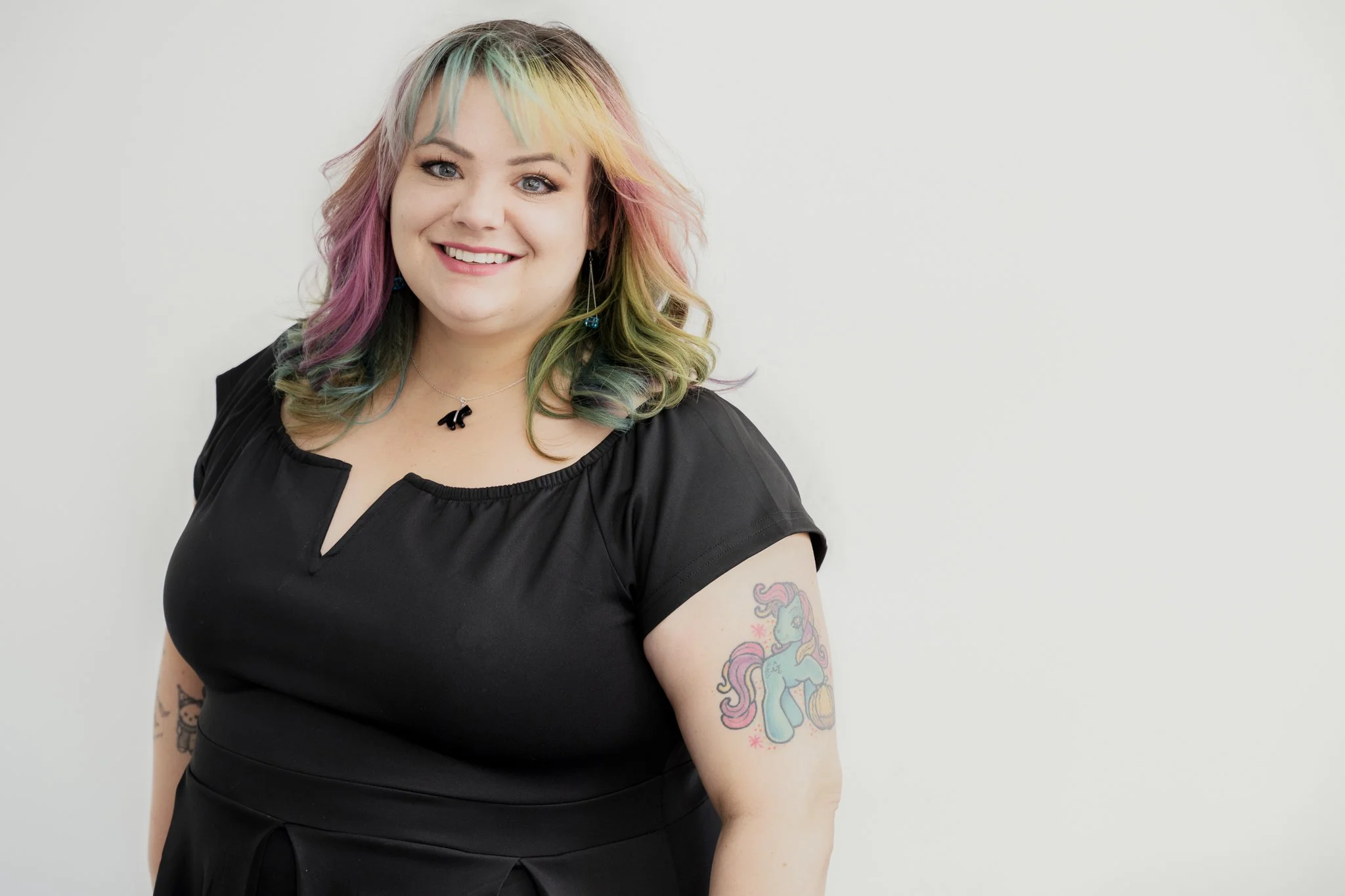 A smiling woman with colorful rainbow hair, wearing a black dress, with tattoos on her arms, standing against a plain white background.
