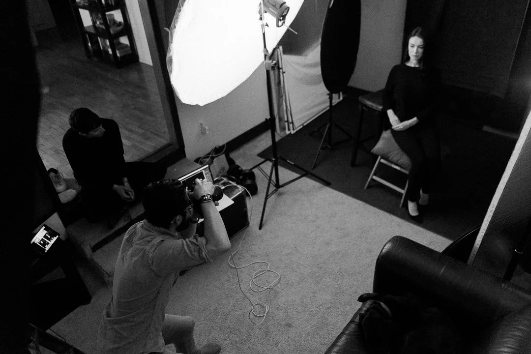 A woman sits on a stool against a black backdrop in a photography studio, while a photographer takes her picture. A large softbox light is directed at her, and a person sits on a bench in the background observing. The studio has various equipment, chairs, and a textured carpeted floor.