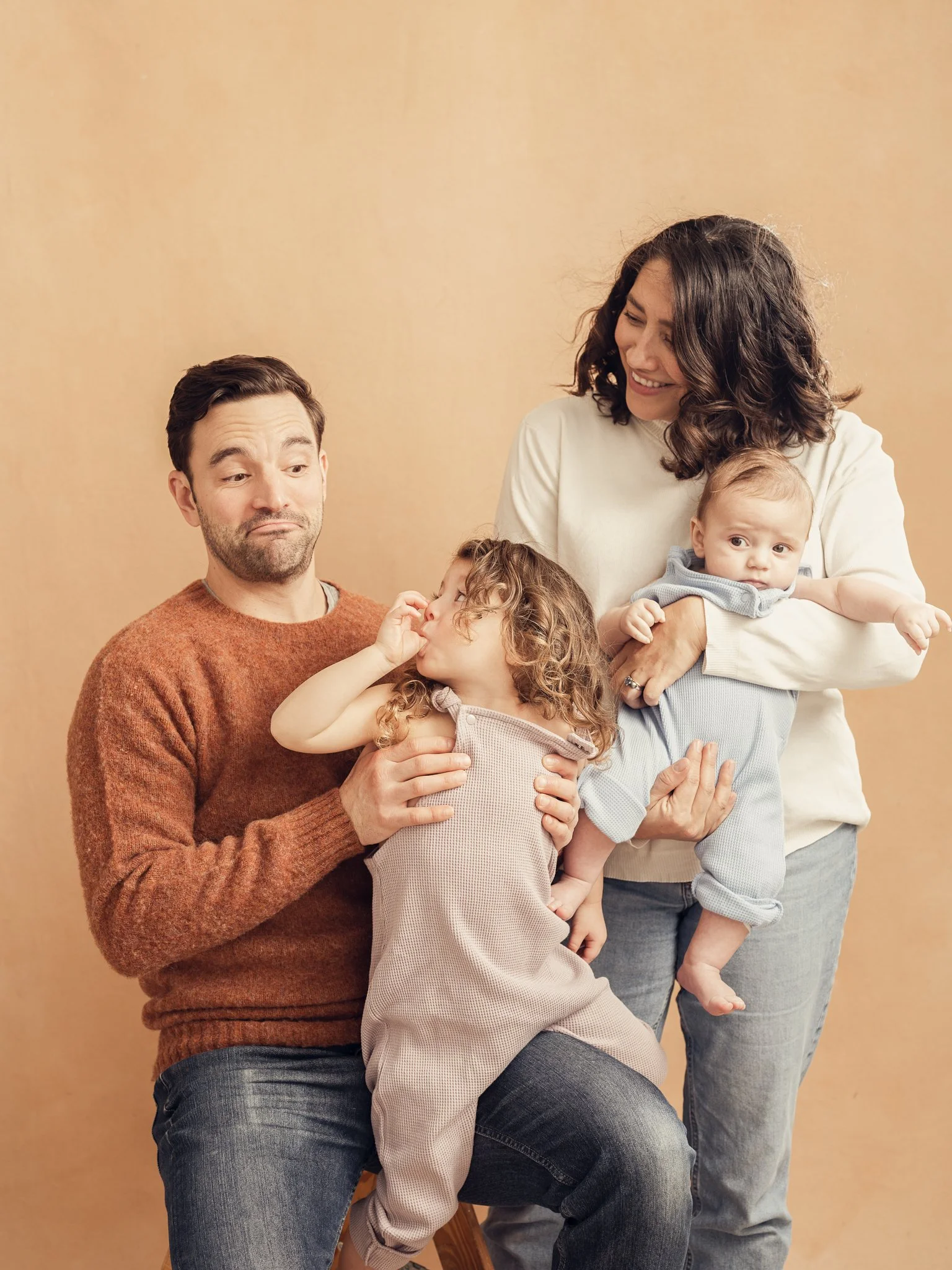 A family of four with two children, a man, a woman, a girl, and a baby, posing together in front of a beige background. The man is sitting, the woman is holding the baby, and the girl is reaching towards the man.