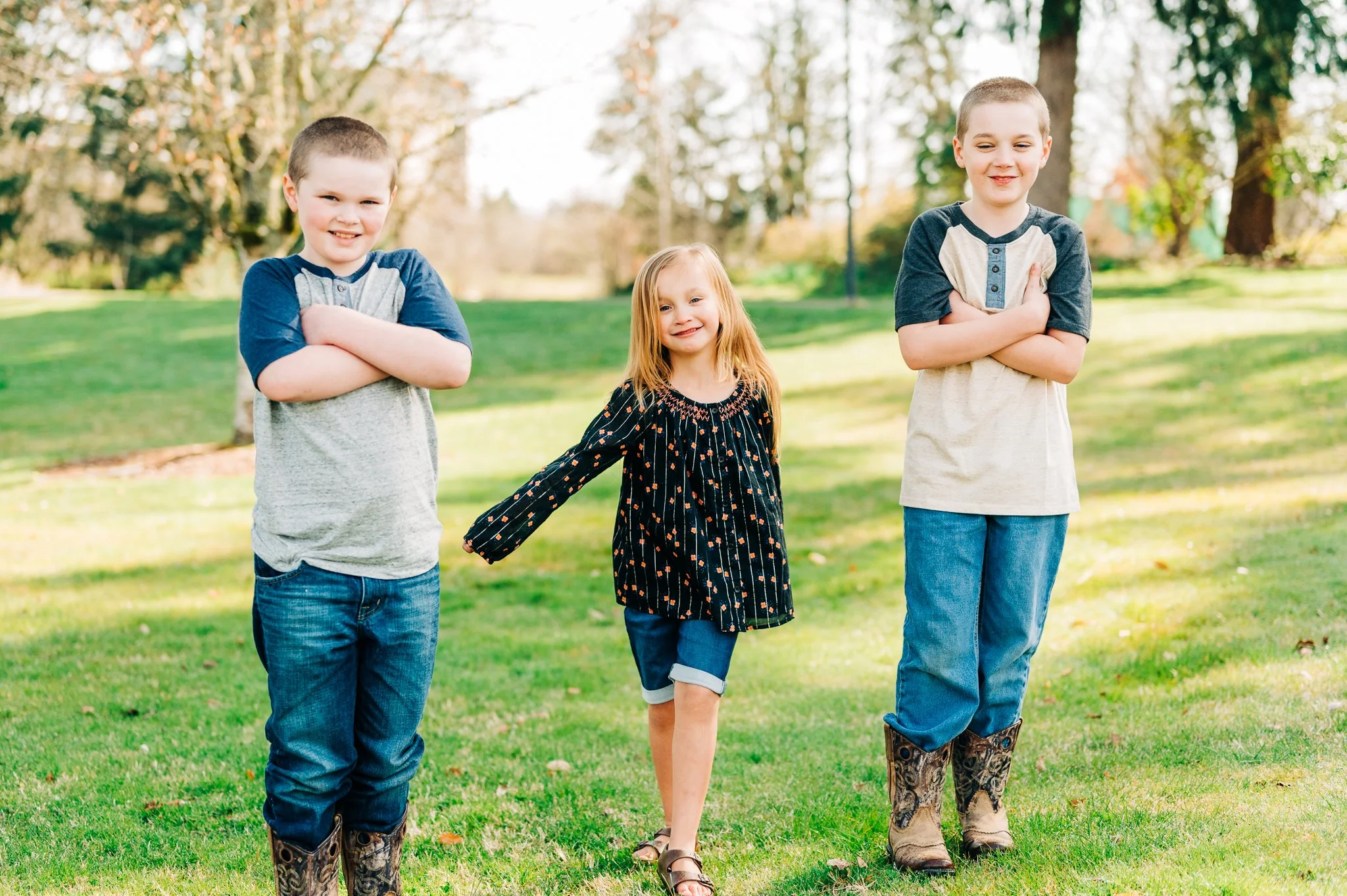 Three children standing in a grassy park with trees in the background, smiling and posing for the camera.