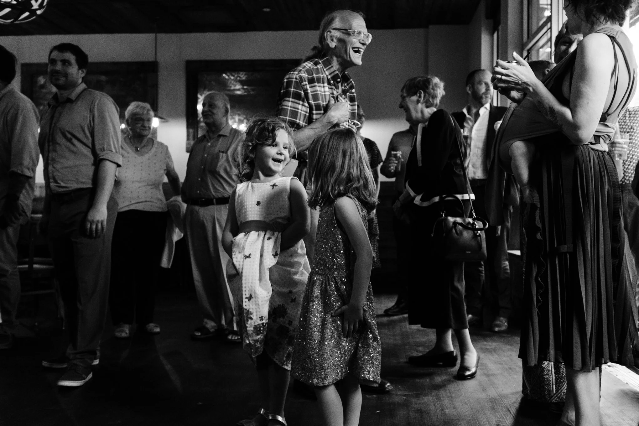 People at a social gathering, including two young girls laughing, an elderly woman smiling, and other adults chatting, in a room with wooden ceiling and framed art on the wall.