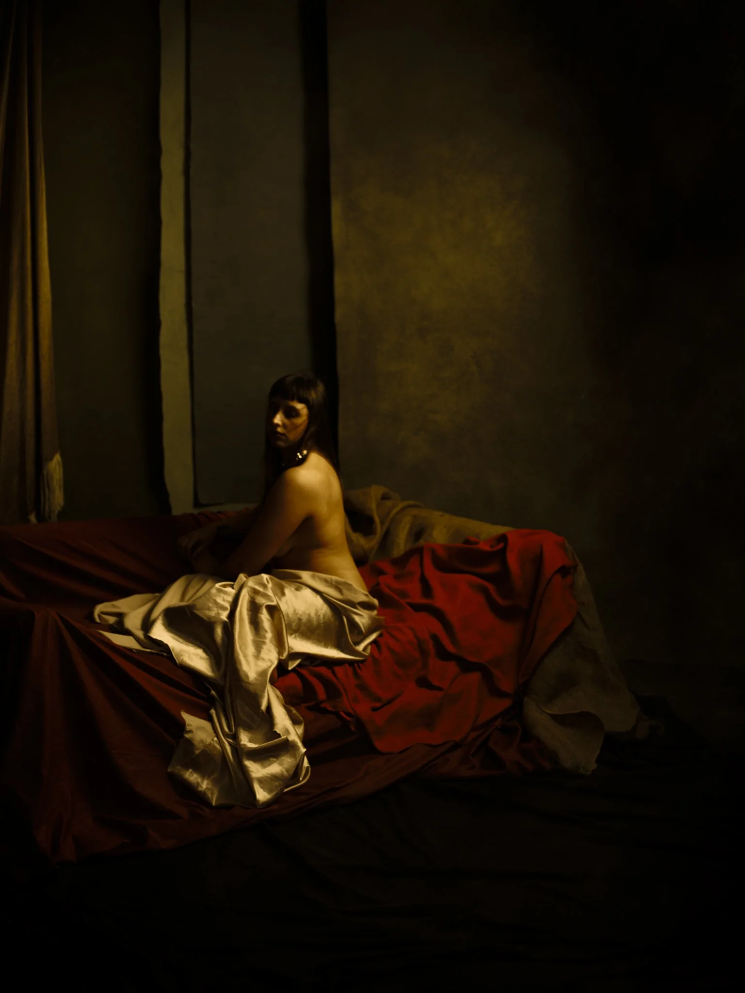 A woman with dark hair sits on a bed with red and gold sheets in a dimly lit room, looking over her shoulder.