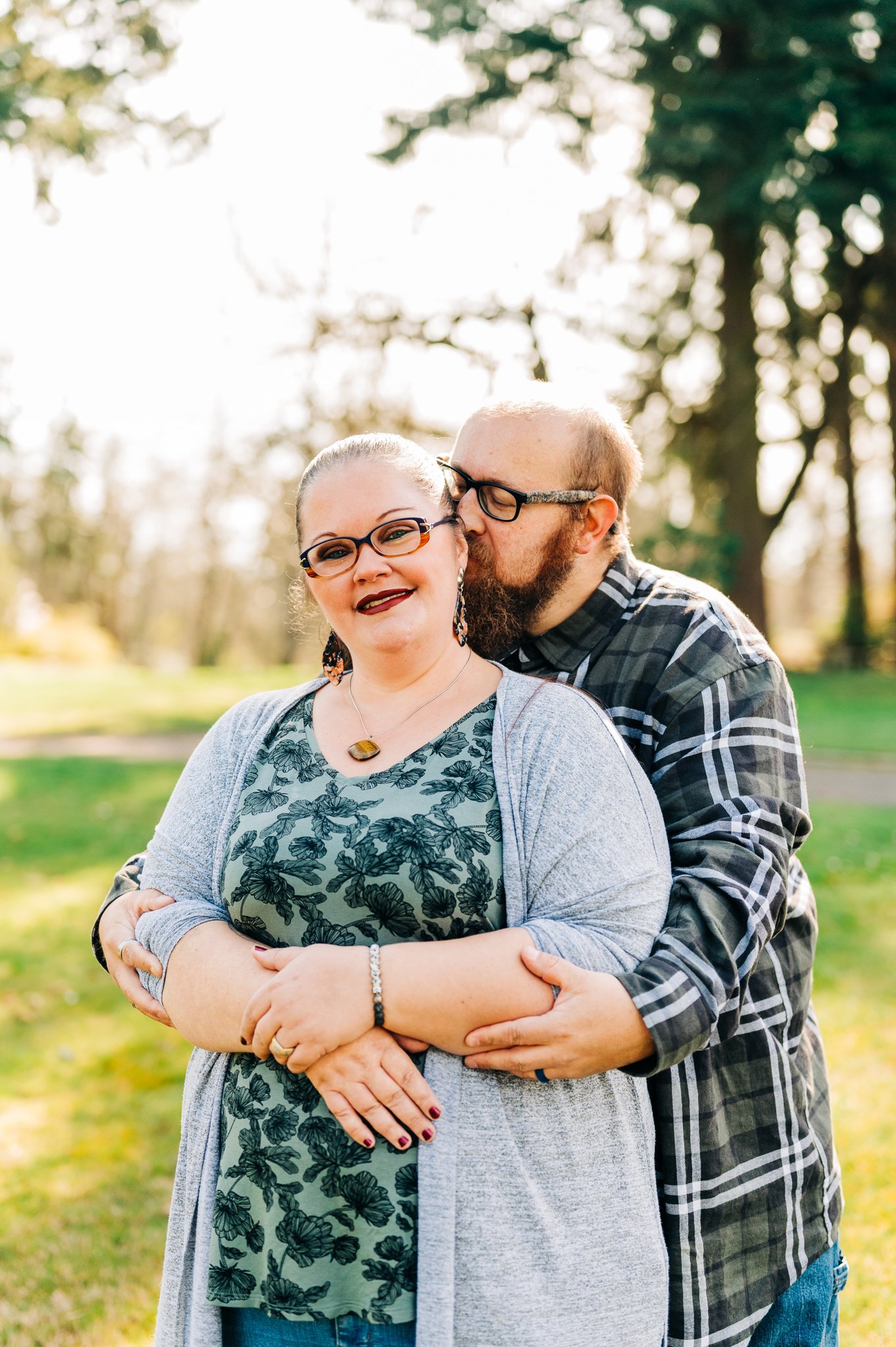 A couple standing outdoors in a park during daytime, with the man kissing the woman on her cheek. They are smiling and appear happy, with trees and sunlight in the background.