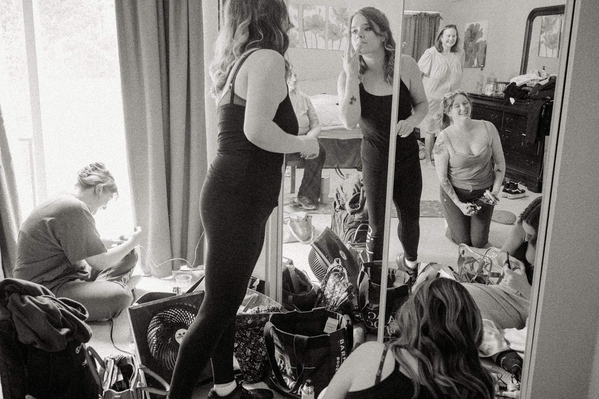 A group of women preparing in a bedroom, some looking at themselves in a mirror, surrounded by clothes and bags.