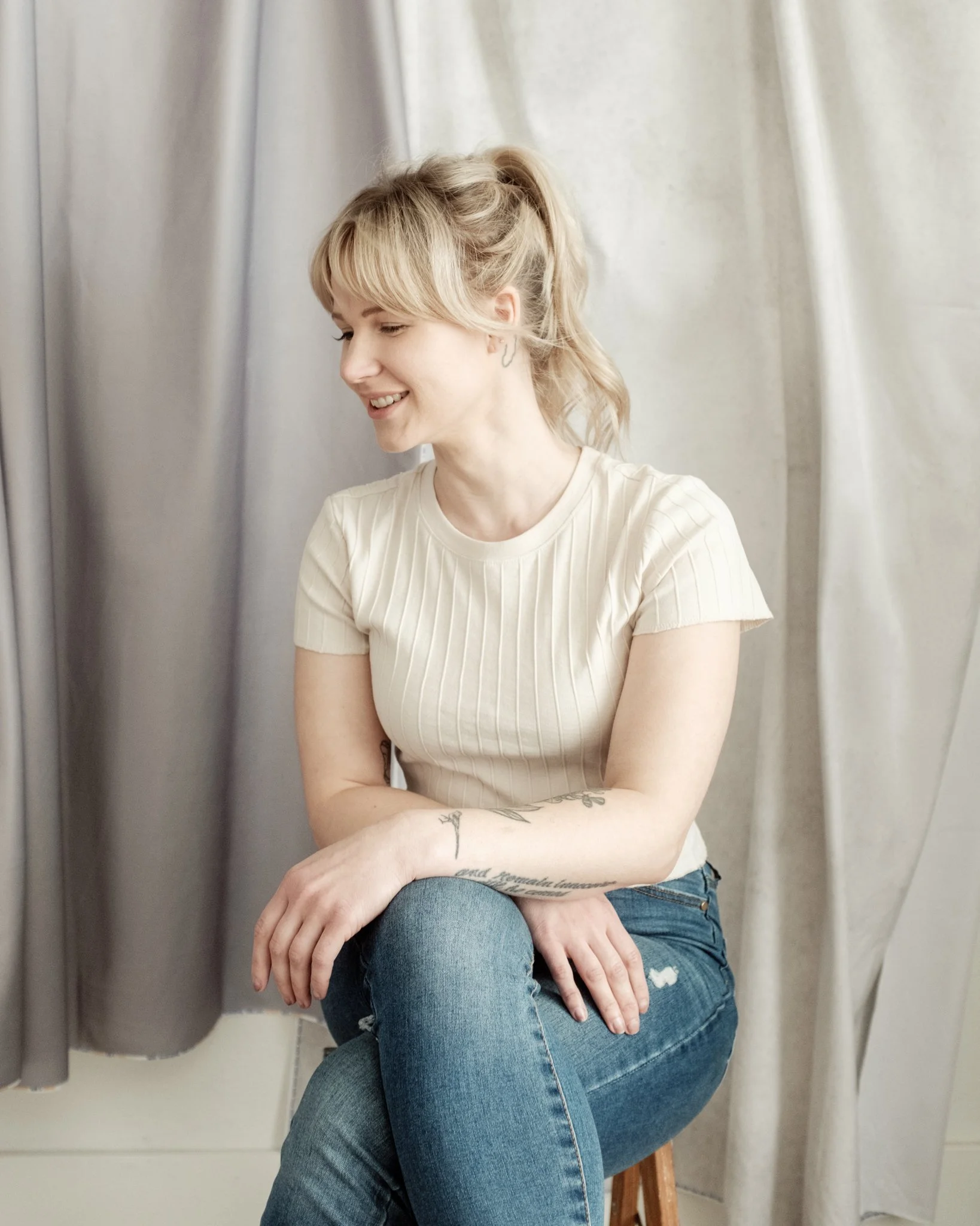 A woman with blonde hair in a ponytail, wearing a cream ribbed t-shirt and blue jeans, sitting on a stool, smiling while looking down. She has tattoos on her left arm.