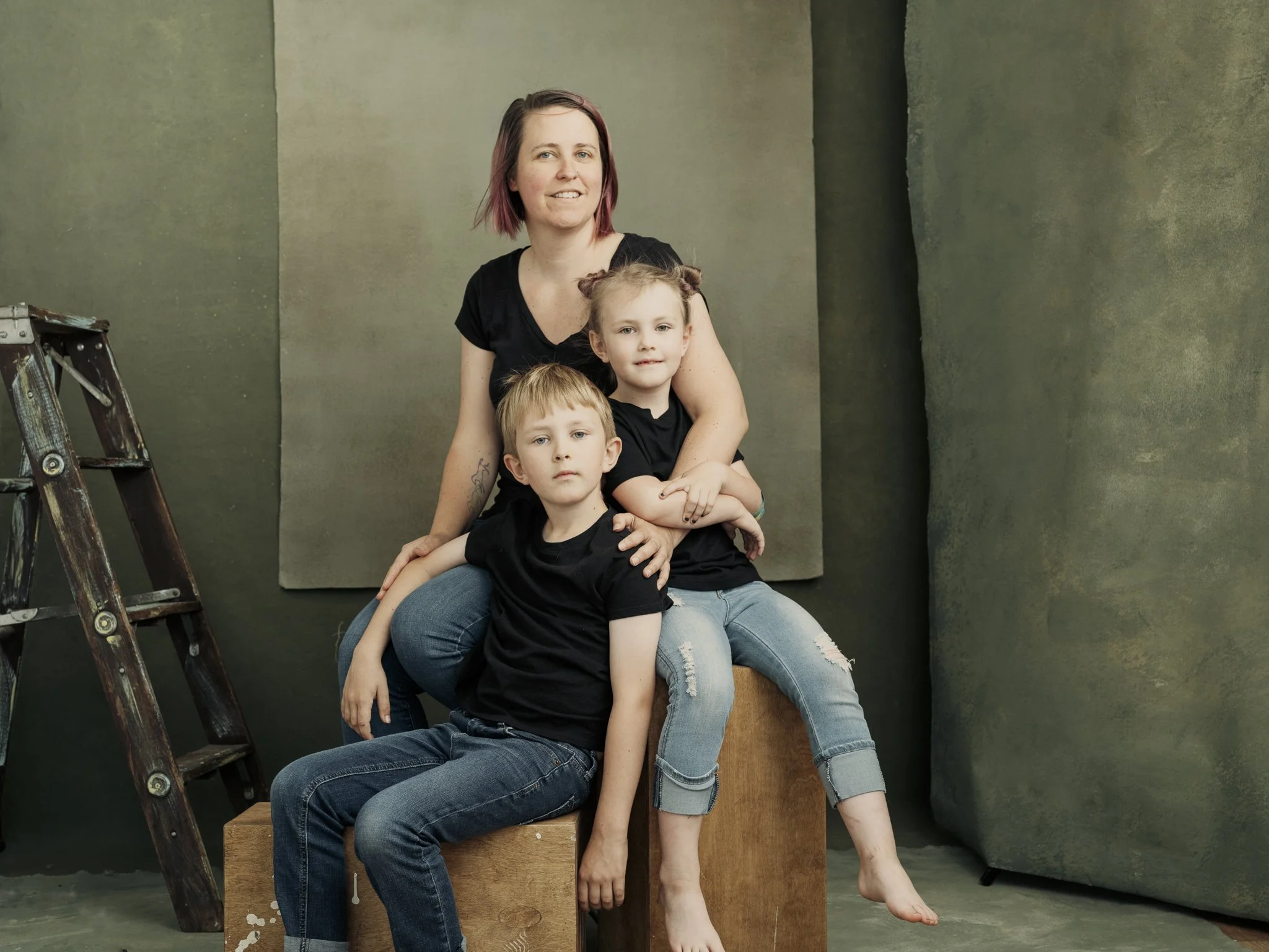 A woman with short hair and two children, a boy and a girl, sitting together in front of a textured green background, with a ladder on the left side.