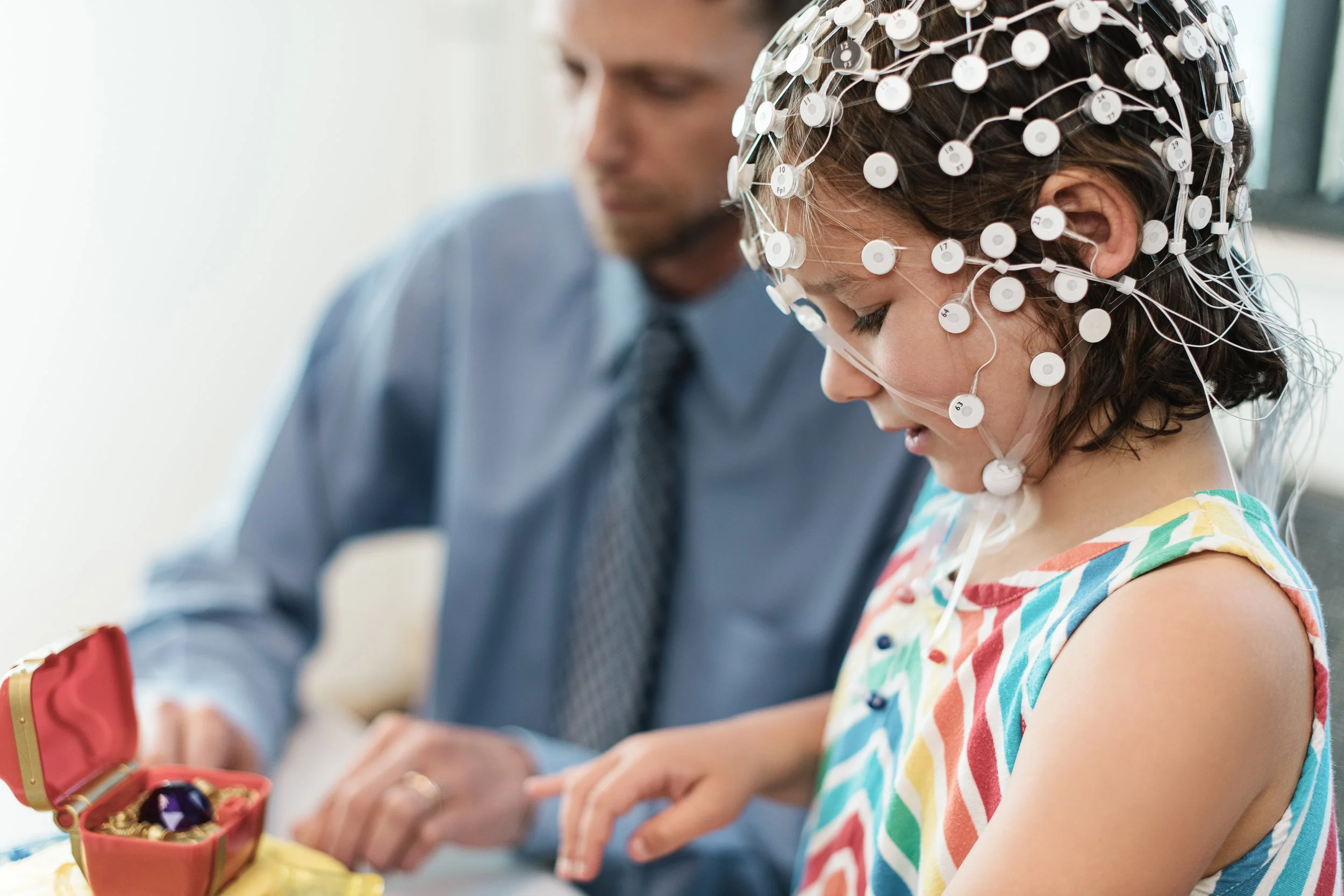 A young girl wearing an EEG cap with numerous electrodes and wires connected to her head, sitting with a woman in a professional setting, possibly a research or medical environment, examining a jewelry box.