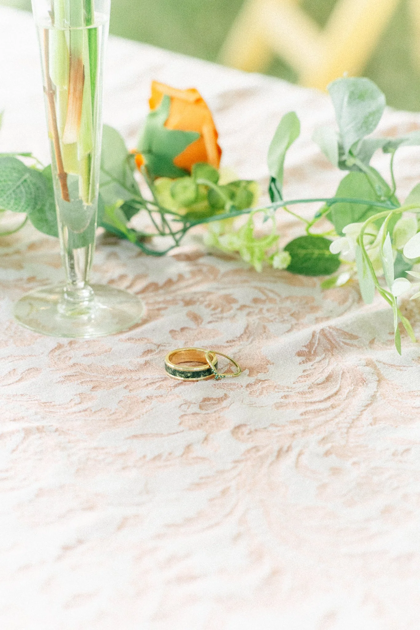 Close-up of a wedding ring lying on a textured tablecloth, with green foliage, a flower, and a glass vase in the background.